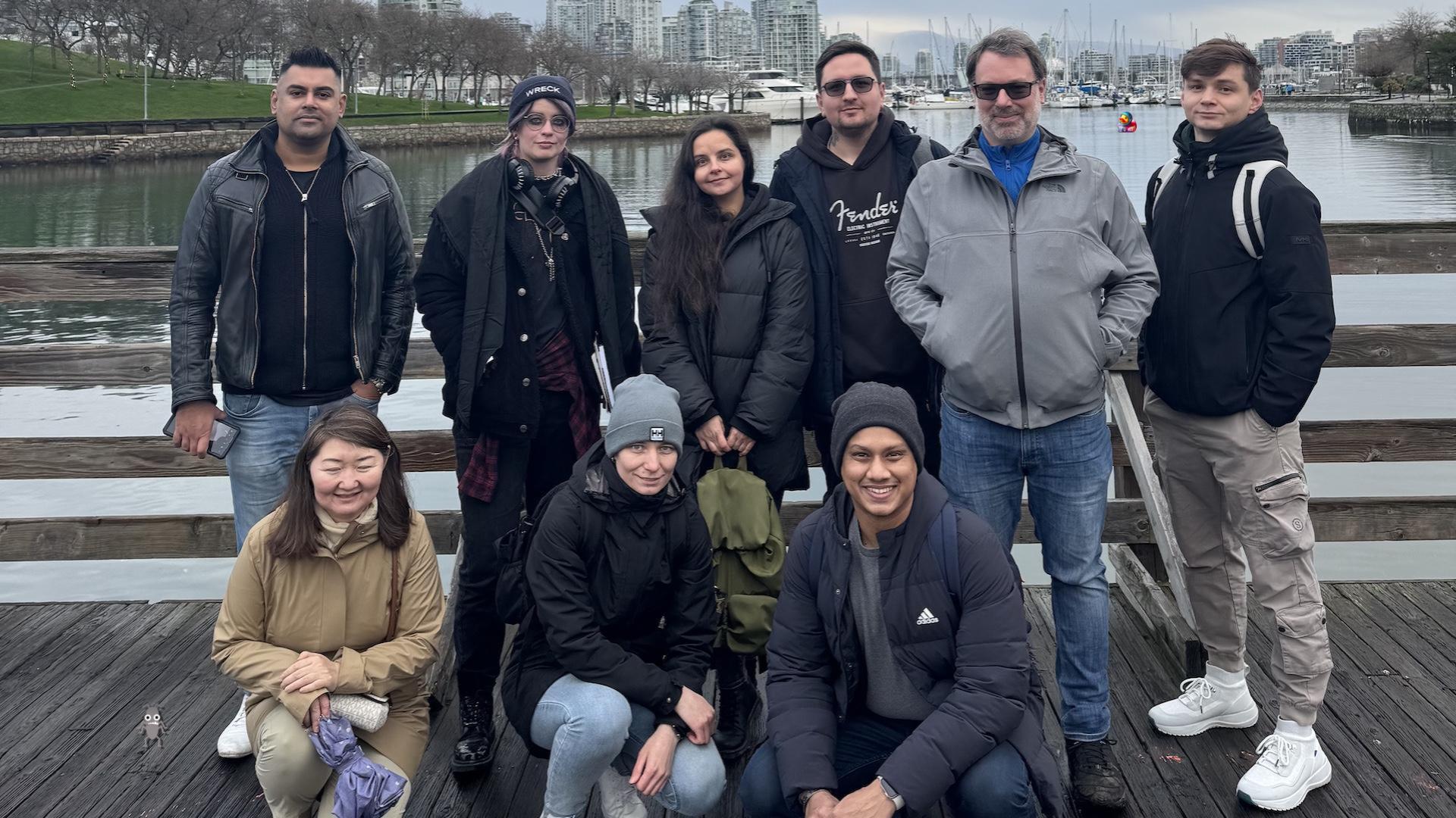 Nine people on a bridge over a river. Six are standing and three are kneeling. Boats, trees and tall buildings are in the background. The sky is a cloudy grey. A duck character and a bug character are attempting to hide in the photo.