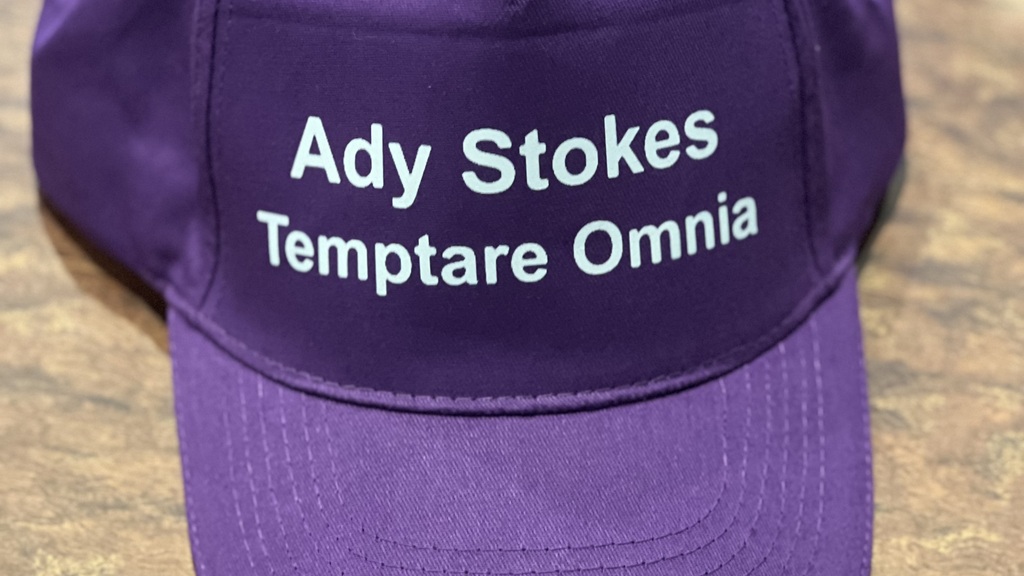 A purple baseball cap on a table top. It says Ady Stokes and Temptare Omnia in white text. 