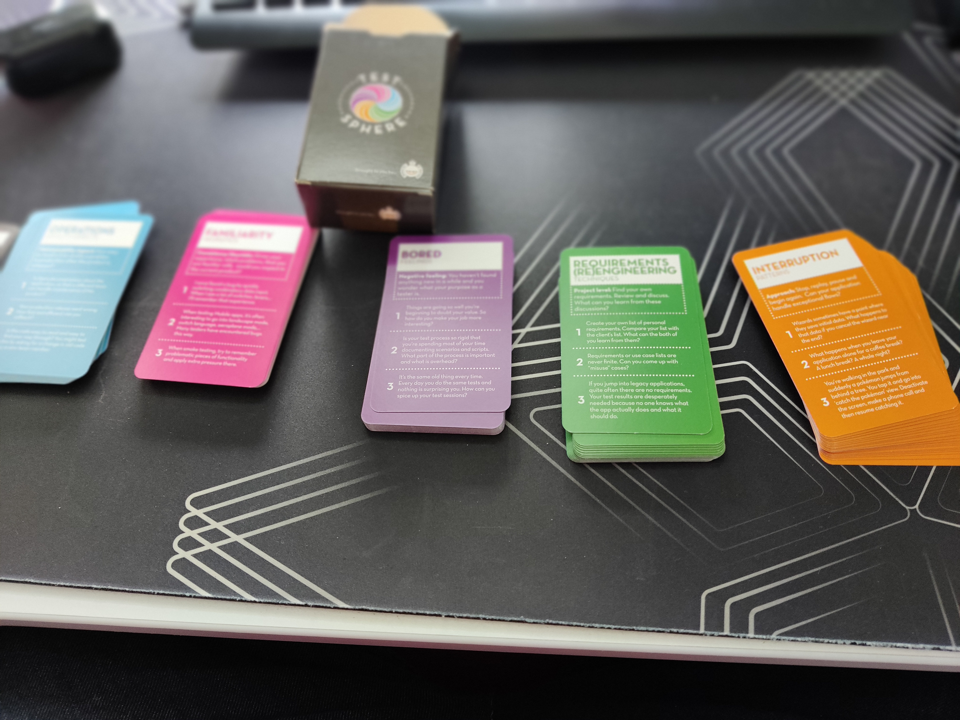 A first-person perspective of a desk setup featuring the TestSphere testing card game. The dark green box stands open behind five colorful cards fanned out horizontally. From left to right, the cards are light blue (Operations), pink (Familiarity), purple (Bored), green (Requirements), and orange (Interruption). Each card contains numbered heuristics and testing prompts. The background shows a blurred keyboard and mousepad.