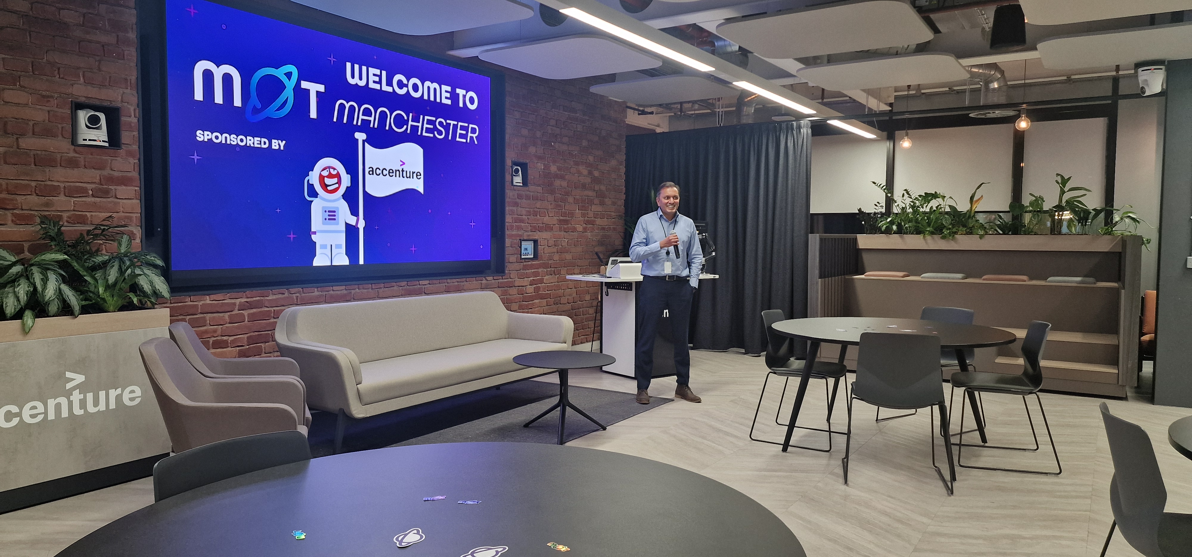 A wide shot of an event space where a person is standing and speaking into a microphone. They are wearing a light blu... image