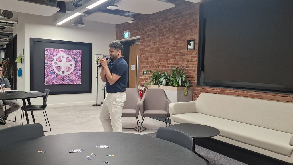 A person stands in the center of a modern office lounge, speaking into a microphone with both hands. They are wearing... image