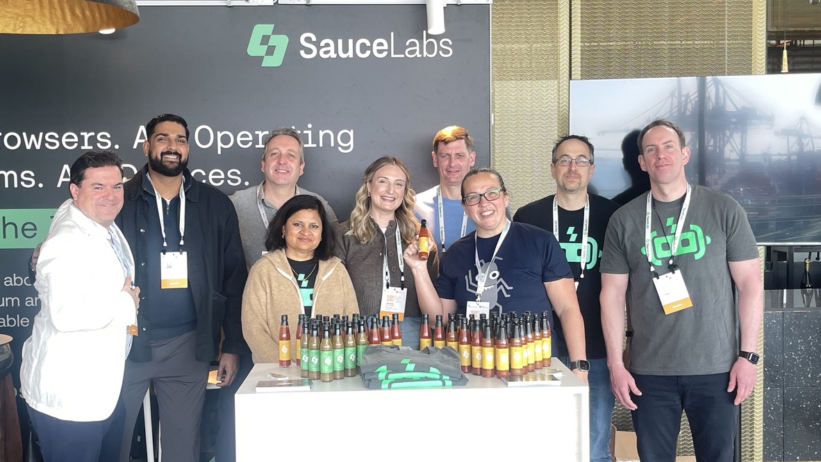 A beaming Rosie stands with the SauceLabs crew behind a stand loaded with bottles of hot sauce and branded tees. Everyone is smiling, and Rosie is proudly holding a bottle like it’s the crown jewel of condiments.