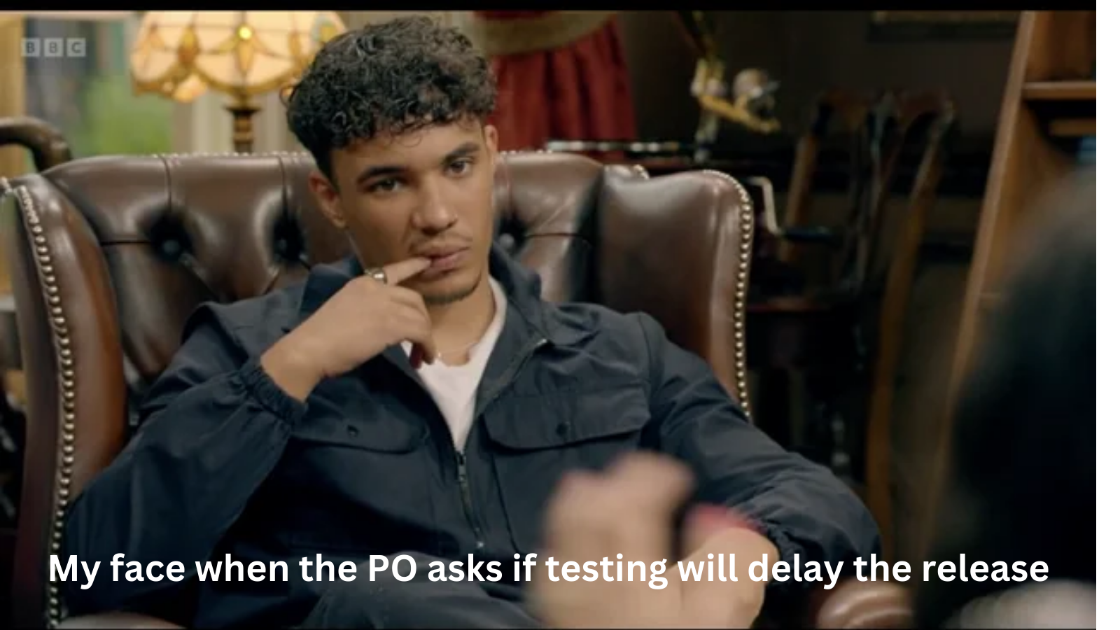 A picture of a young man biting his finger, while sat on a leather chair an annoyed look on his face, with text reading "My face when the PO asks if testing will delay the release."