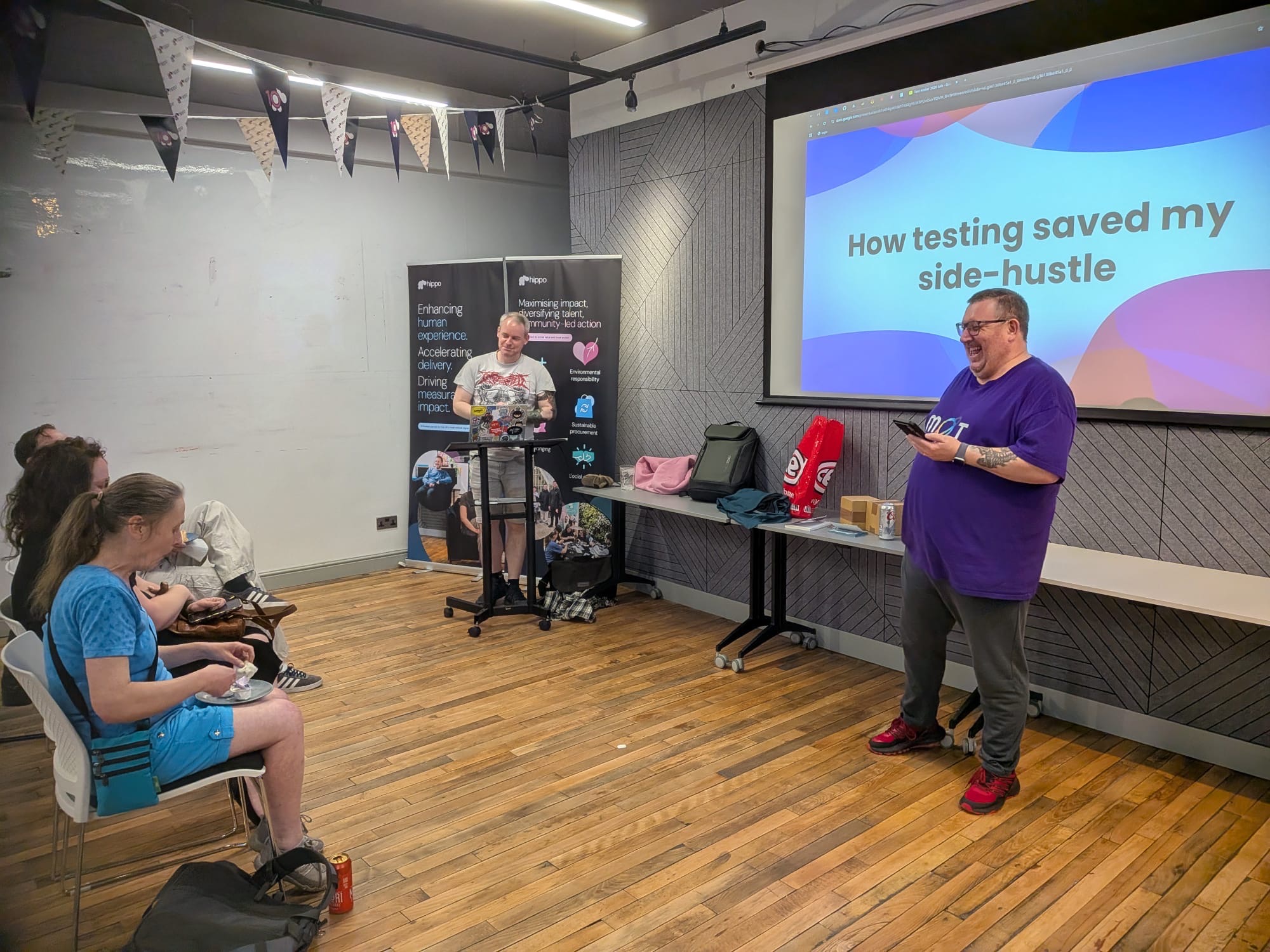 Ady is stood at the front laughing in his purple MoT rainbow T-shirt. Colin is at the podium with his laptop and in g... image