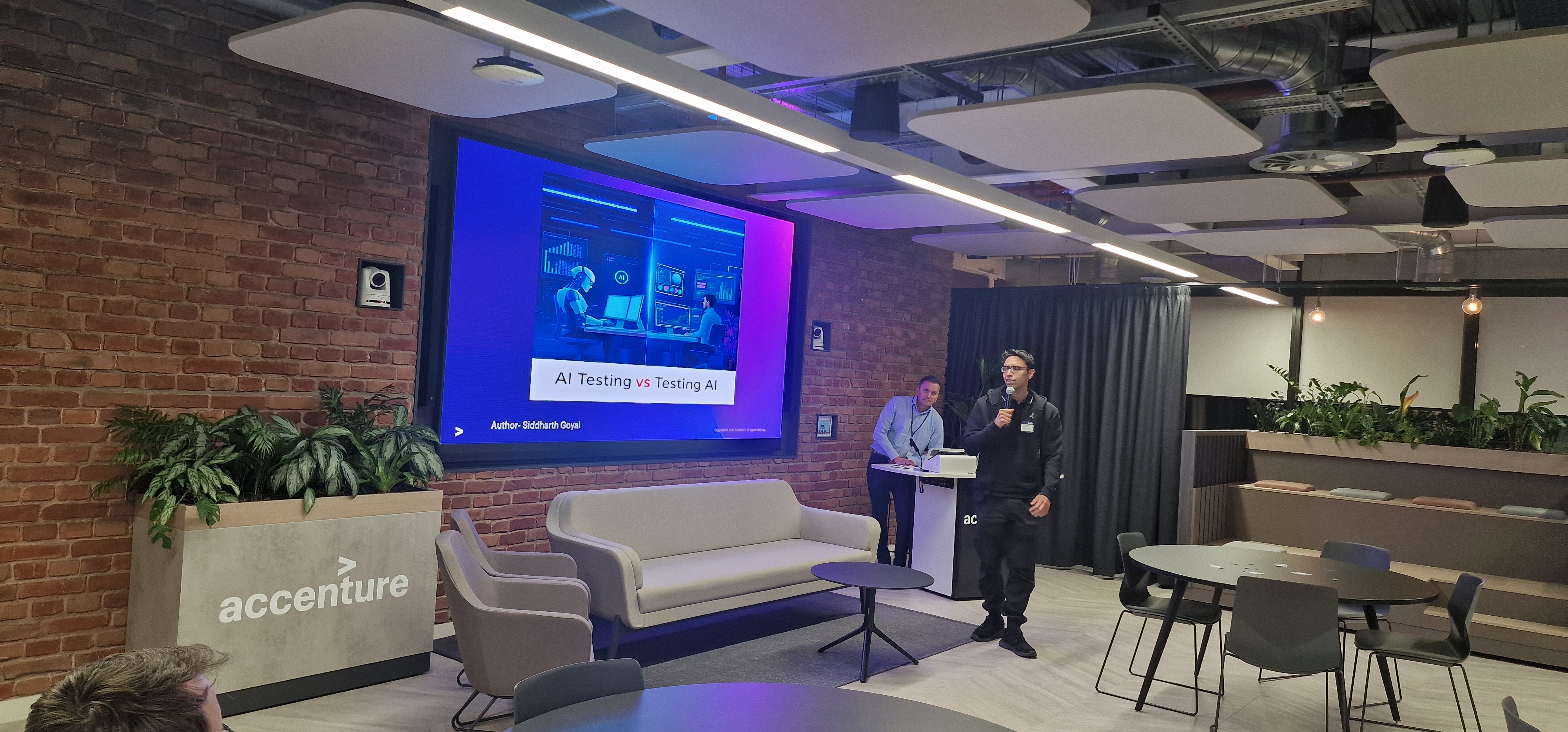A speaker stands in a modern event space, holding a microphone and addressing an audience. He is wearing a black hood... image