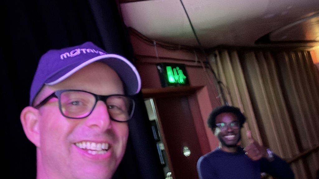 A smiling man takes a selfie, wearing a purple "MoTaverse" hat and lanyard, within a theatre space. His colleague sta... image
