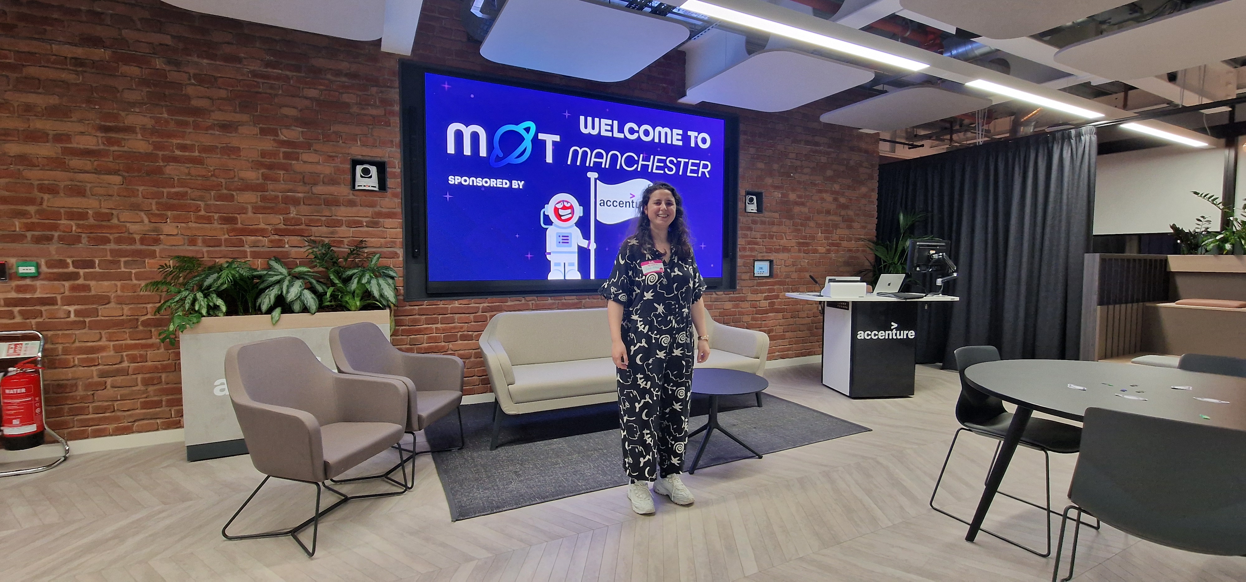 A person stands smiling in a modern office lounge, wearing a dark short-sleeved jumpsuit with a white abstract patter... image