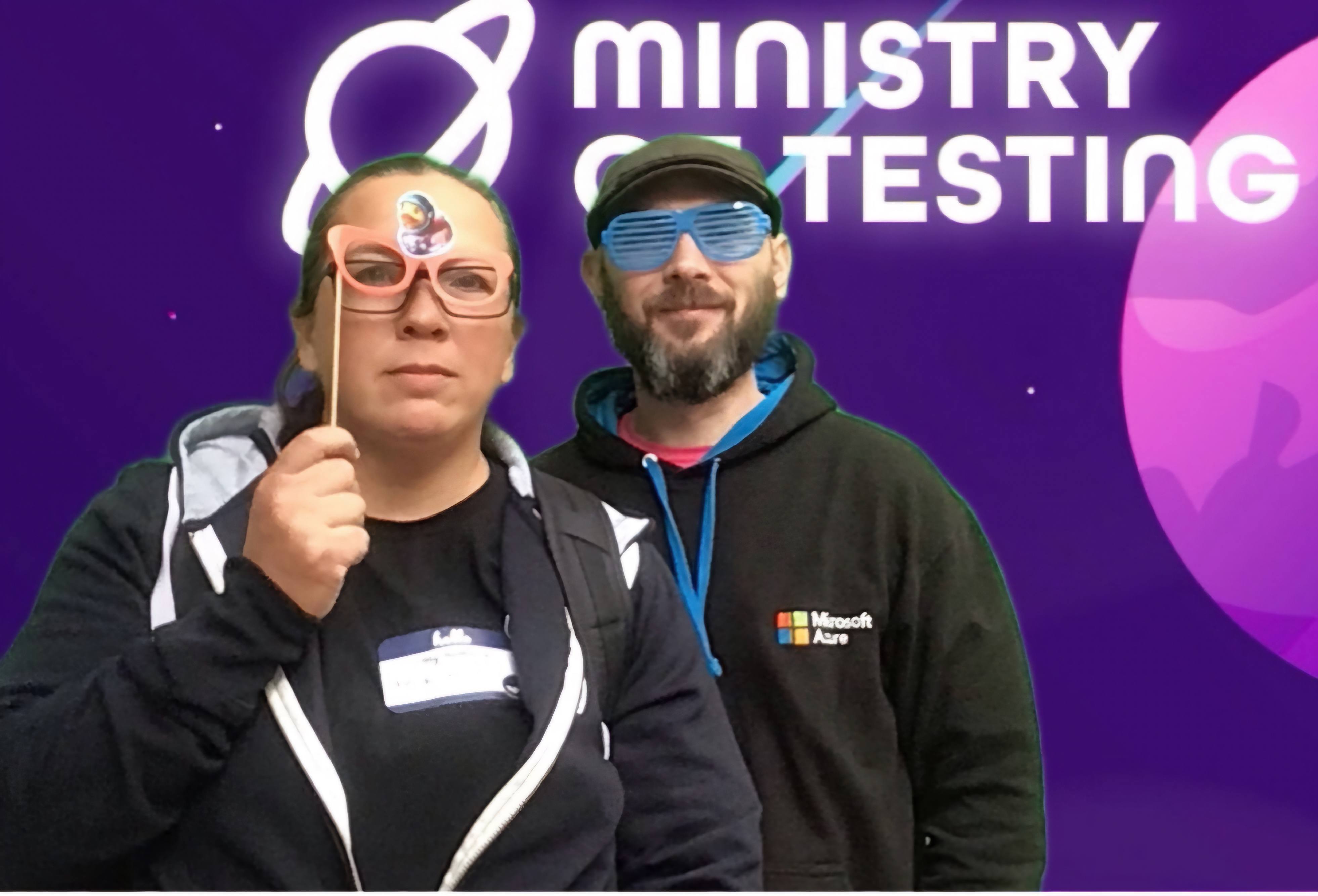 Two people posing in front of a Ministry of Testing backdrop. The person on the left holds up a paper prop to their face, while the person on the right wears colourful sunglasses. They are both smiling and wearing hooded sweatshirts.