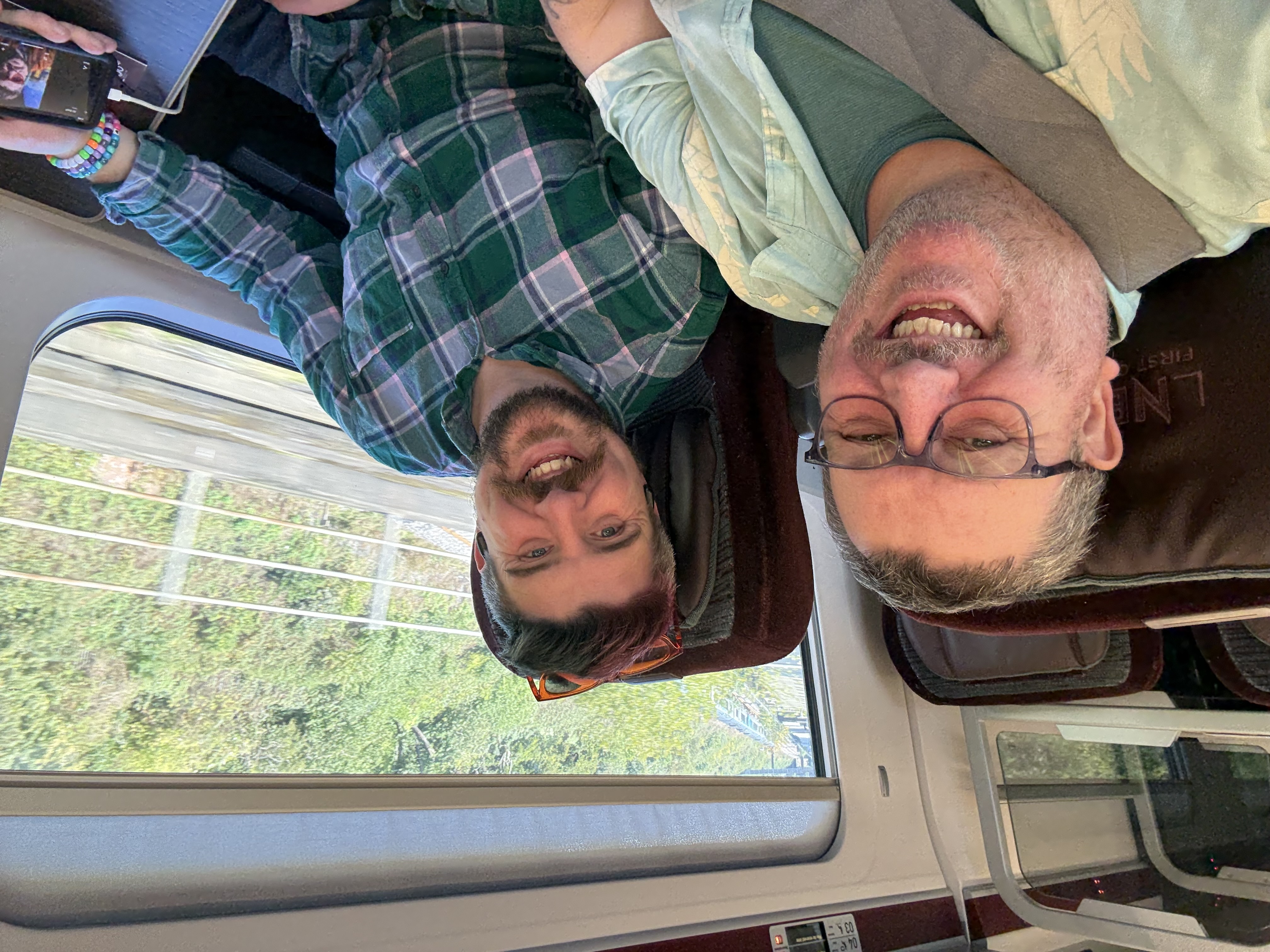 Scott and Ady sat in first class. Scott has pink and blue hair and a green checked shirt. Ady has glasses and is wearing a blue Hawaiian style shirt over a green tshirt