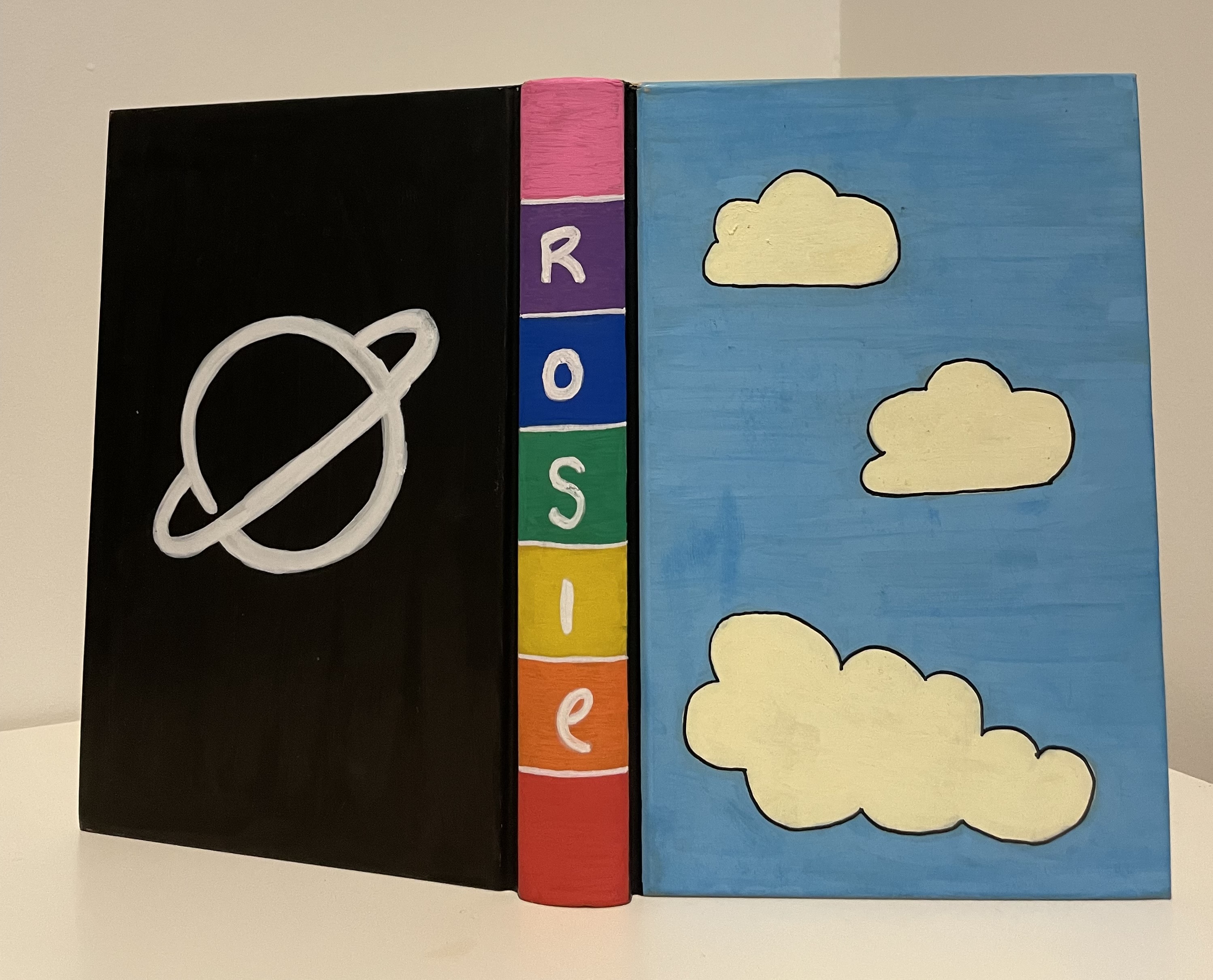 A hard back notebook painted by myself with paint pens.

The back cover is black with a MoT planet logo. The binding says Rosie with rainbow colours in the background. The front has a blue sky and 3 white clouds.