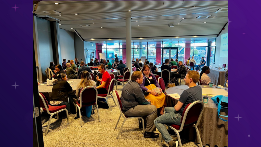 A medium shot of an indoor conference room shows people seated around round tables. Some are facing the camera, while others are seen from the back. The space is well-lit with large windows and modern décor. A screen is visible in the background, displaying a presentation.
