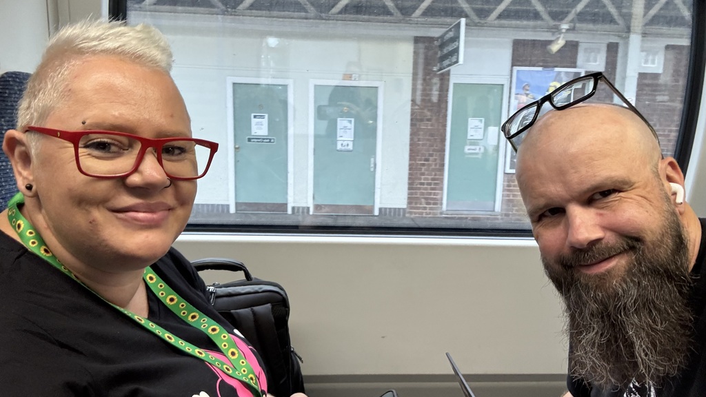 Tamara (Tami) and Christian smiling for a selfie while sitting together on a train. Tami has short blonde hair and re... image