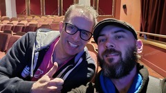 Two people smile for a selfie in a theatre with red seats and decorative walls. One person gives a thumbs up, wearing glasses and a hoodie. The other person wears a cap and has a beard. image
