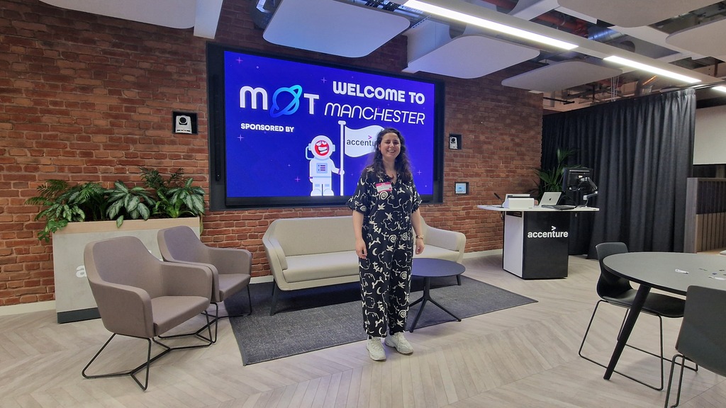 A person stands smiling in a modern office lounge, wearing a dark short-sleeved jumpsuit with a white abstract patter... image