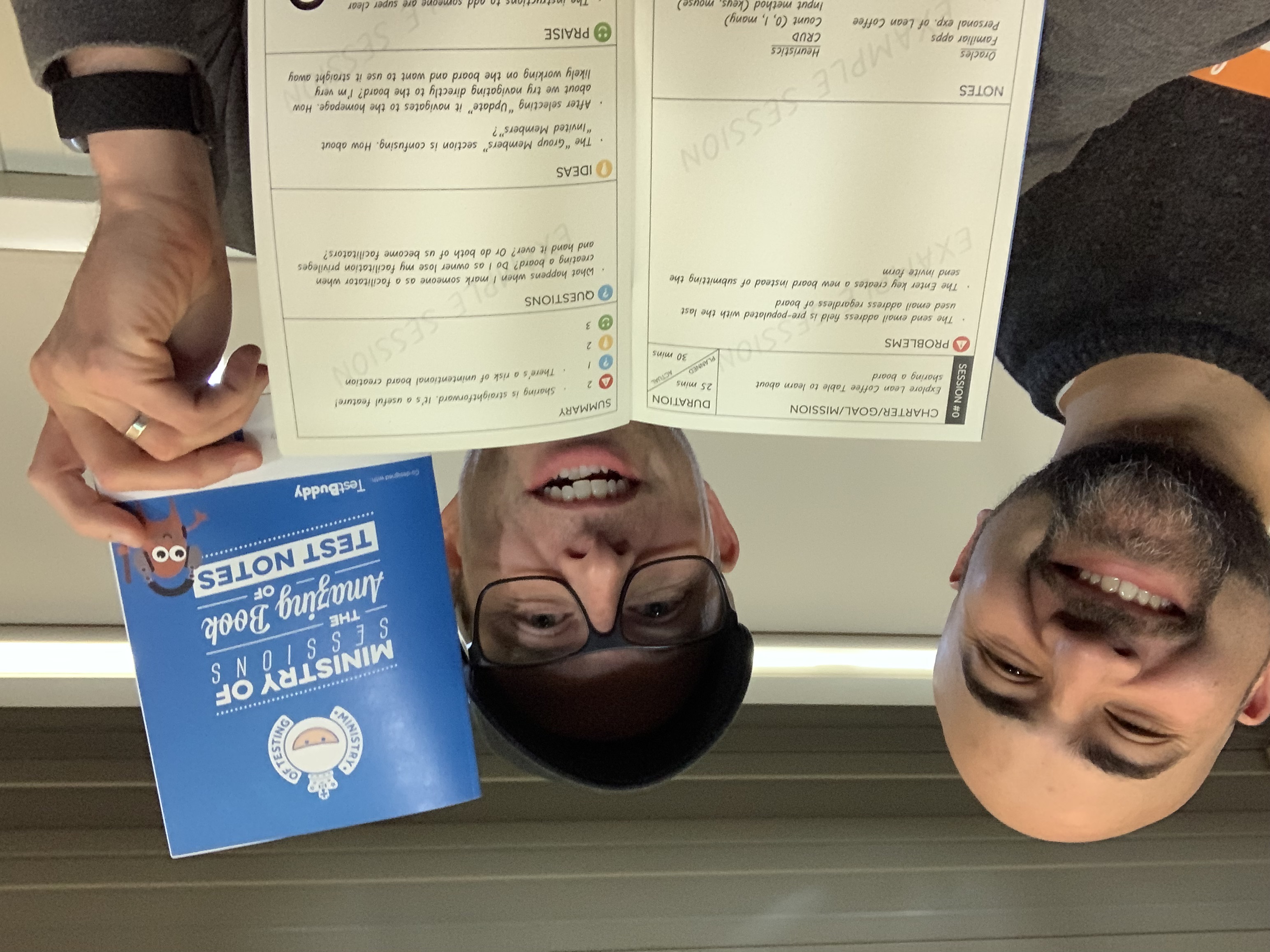 Two men, looking very happy holding up a physical notebook with the following words on the front cover: Ministry of Sessions: The Amazing Book of Test Notes
