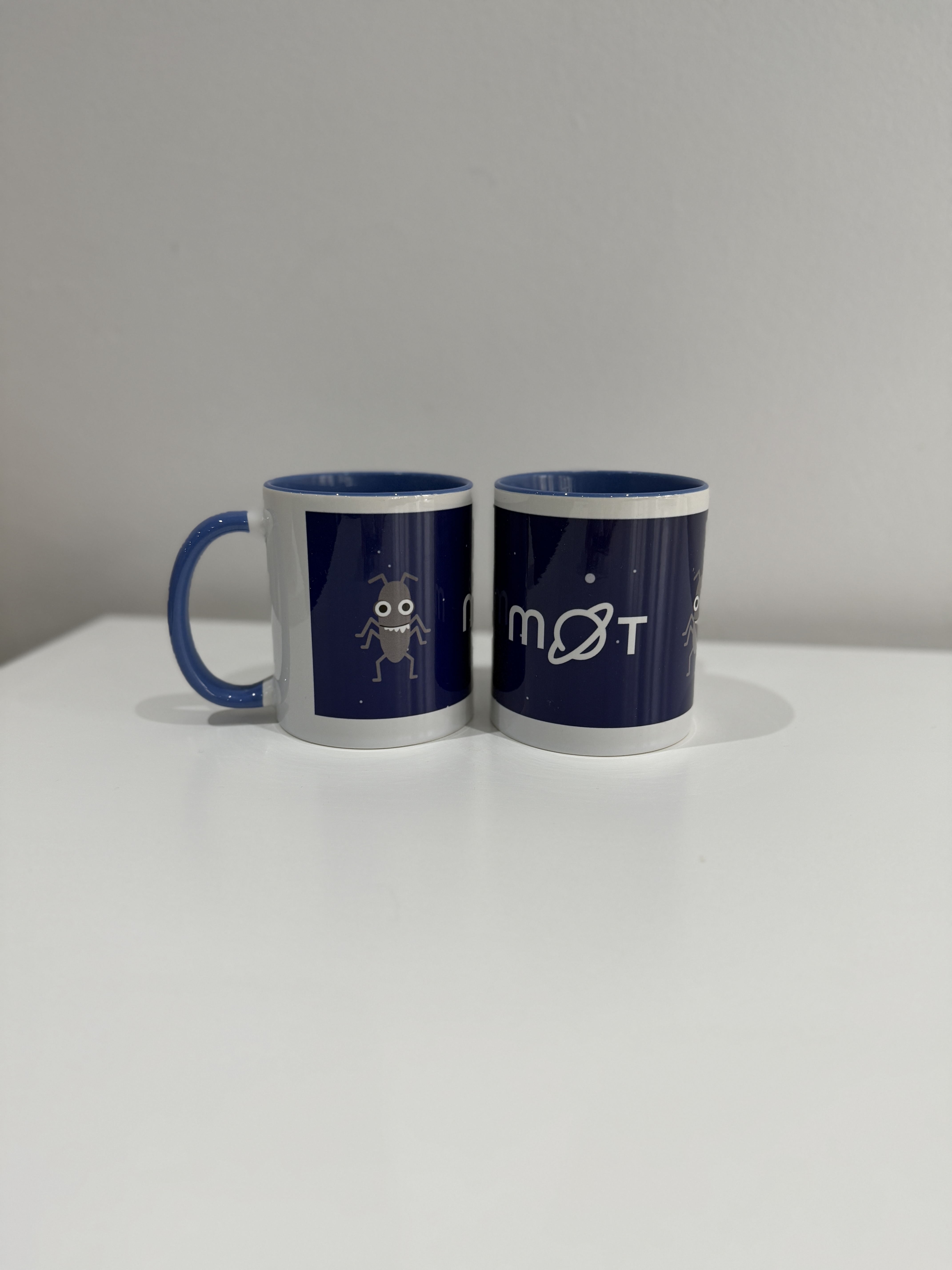 Two mugs with bug characters on them. There is the MoT logo on a starry purple background. The mug on the right appears to be missing a handle. 