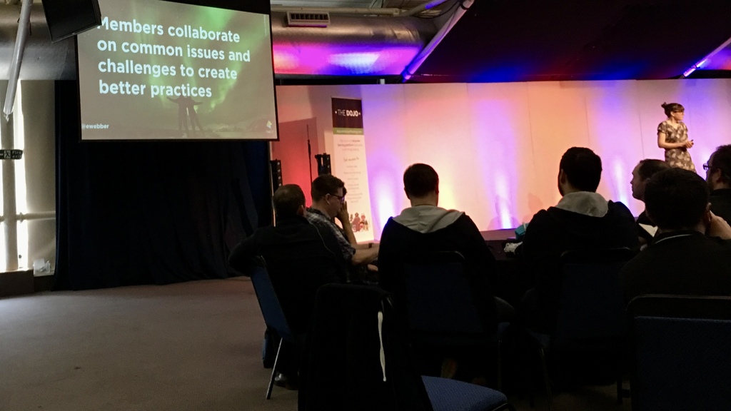 A woman on stage in a large event space. The slide on the screen reads: Members collaborate on common issues and chal... image
