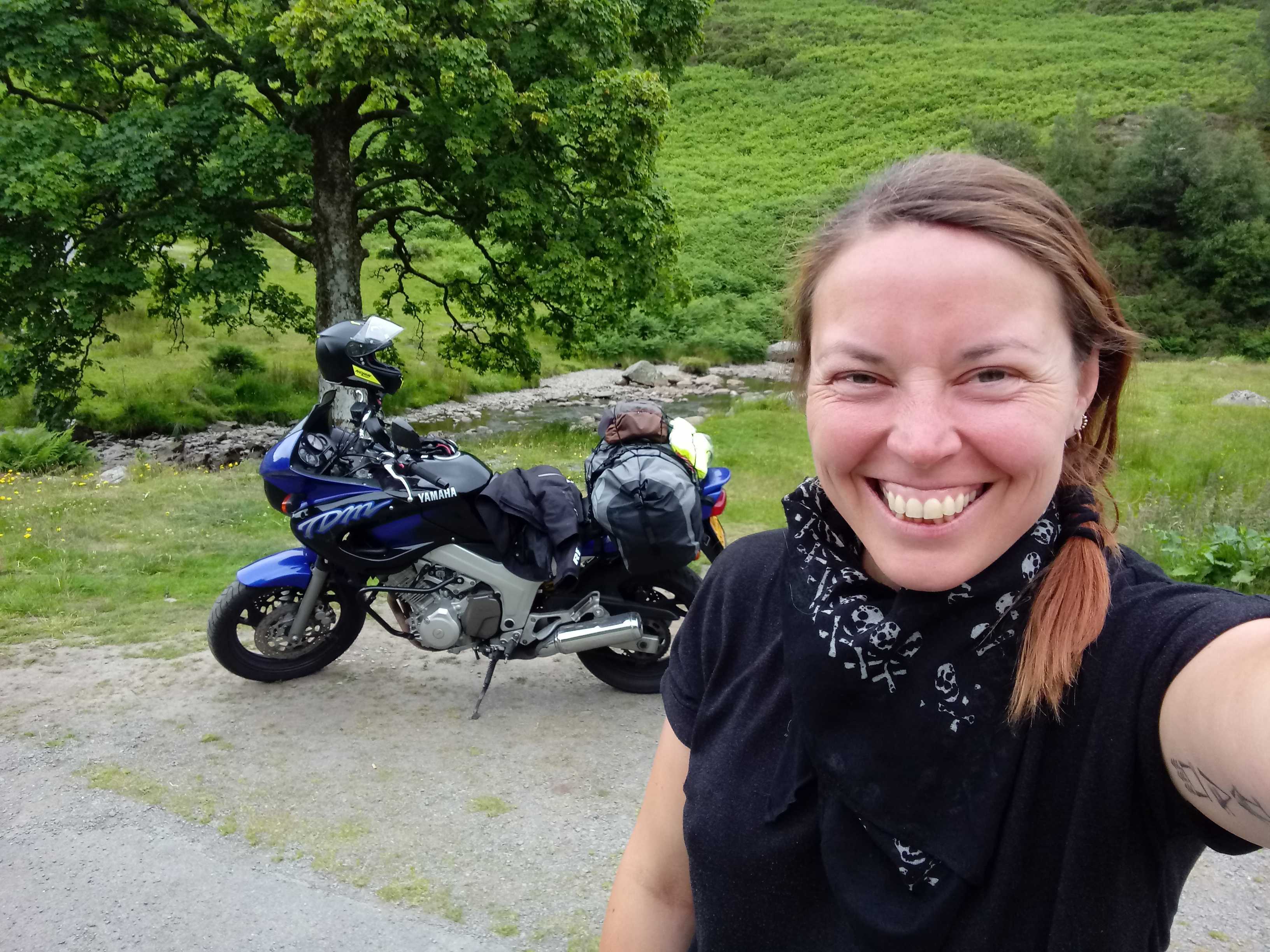 Me, a white woman with medium length brown hair in a plait, looking very excited in front of my black and blue Yamaha motorbike, in a green Welsh landscape, near a large tree and a river