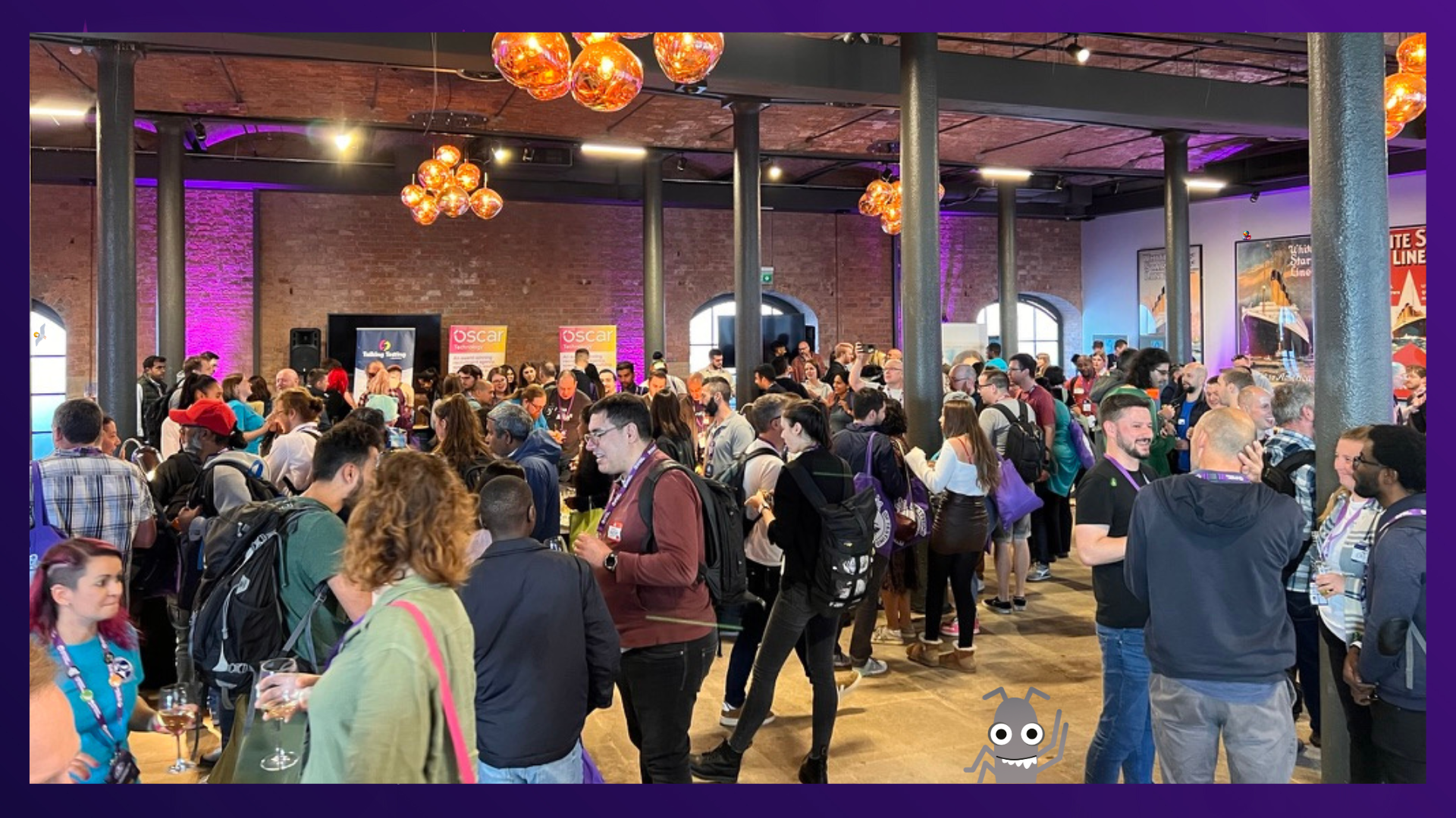 A photograph shows a large group of people gathered in a spacious, high-ceilinged room with exposed brick pillars. The room has a modern, industrial feel, and is lit by warm, ambient lighting from several unique, spherical light fixtures hanging from the ceiling. A large crowd of people, many holding drinks, are mingling and talking in small groups. Some are wearing lanyards with what appears to be a conference or event logo. In the foreground, people are seen from the chest up, some with their backs to the camera, while in the background, a stage setup with what looks like a screen is visible. The atmosphere appears social and lively. A cartoon bug with large eyes and antennae is superimposed on the bottom of the image, appearing as if it's on the floor looking up at the people. There is also a cartoon seagull and duck in the image. 