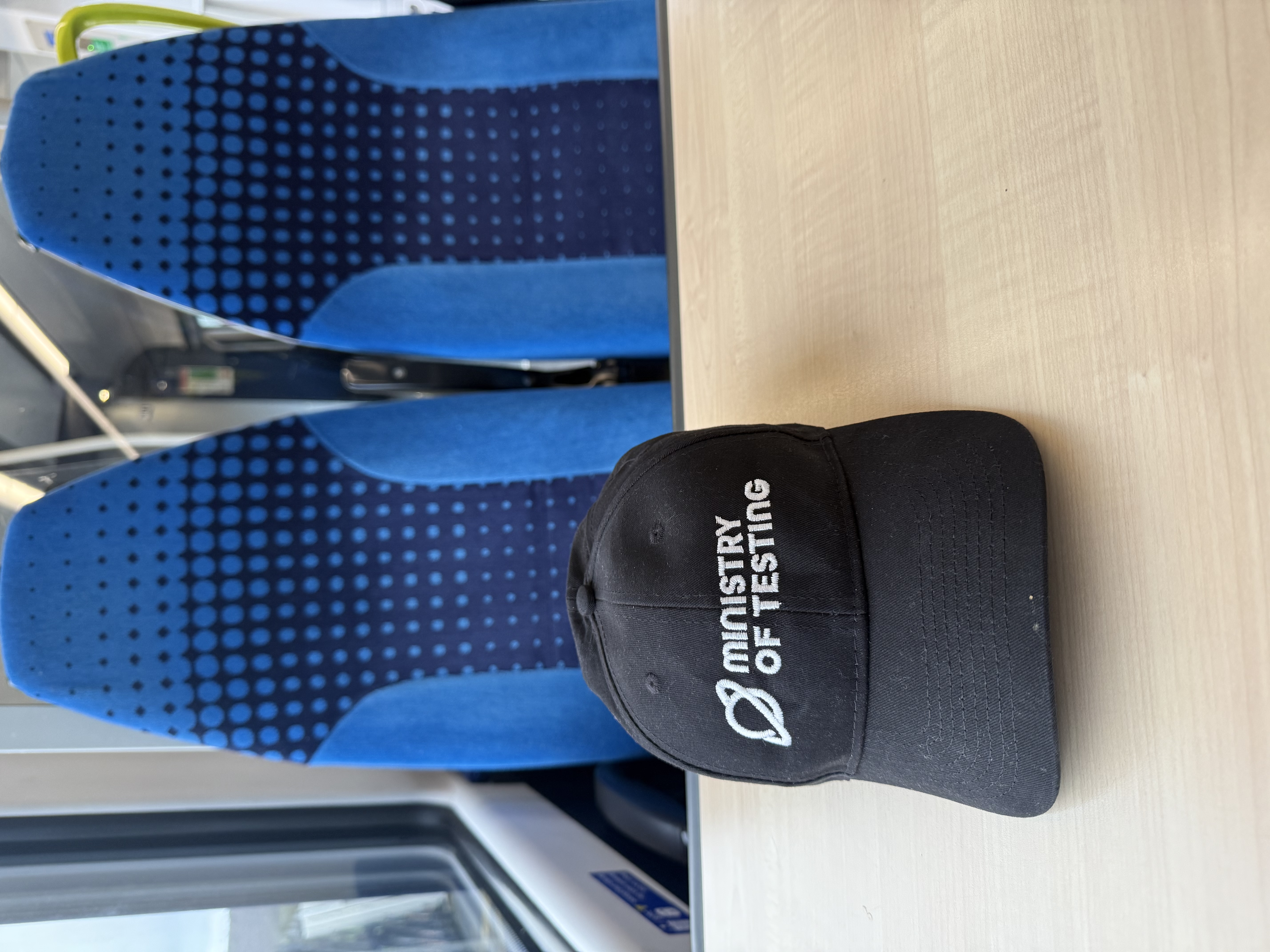 A black baseball cap with the planet logo and Ministry of Testing sits on a train table with two blue train seats in the background with a spotted pattern 