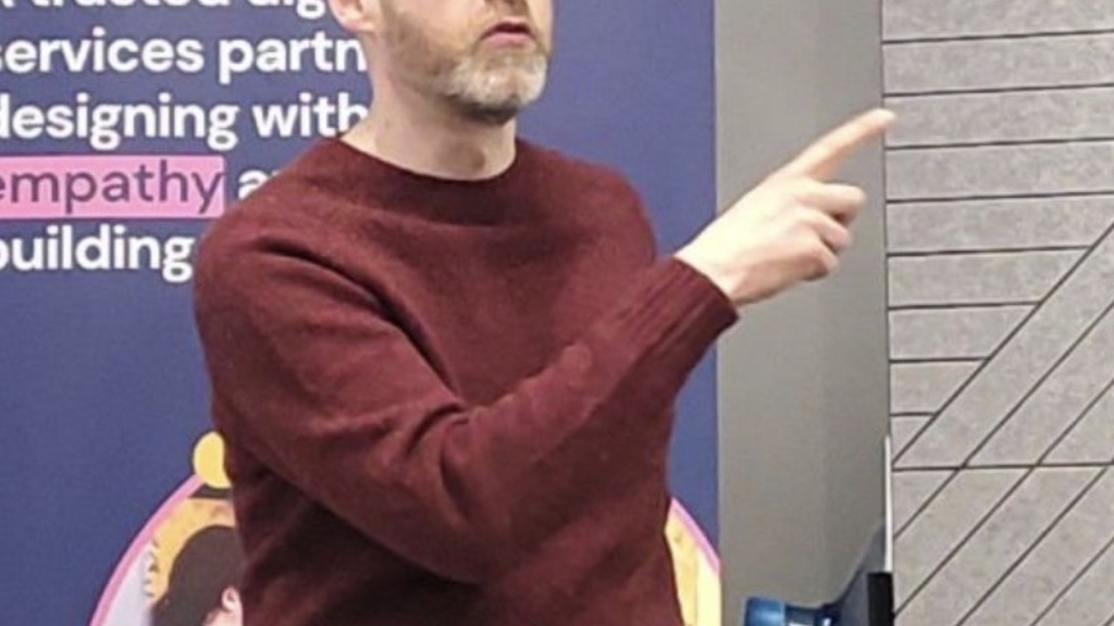 Alex is a white male in black jeans and a plum top, wearing glasses. He is stood speaking and pointing in front of a ... image