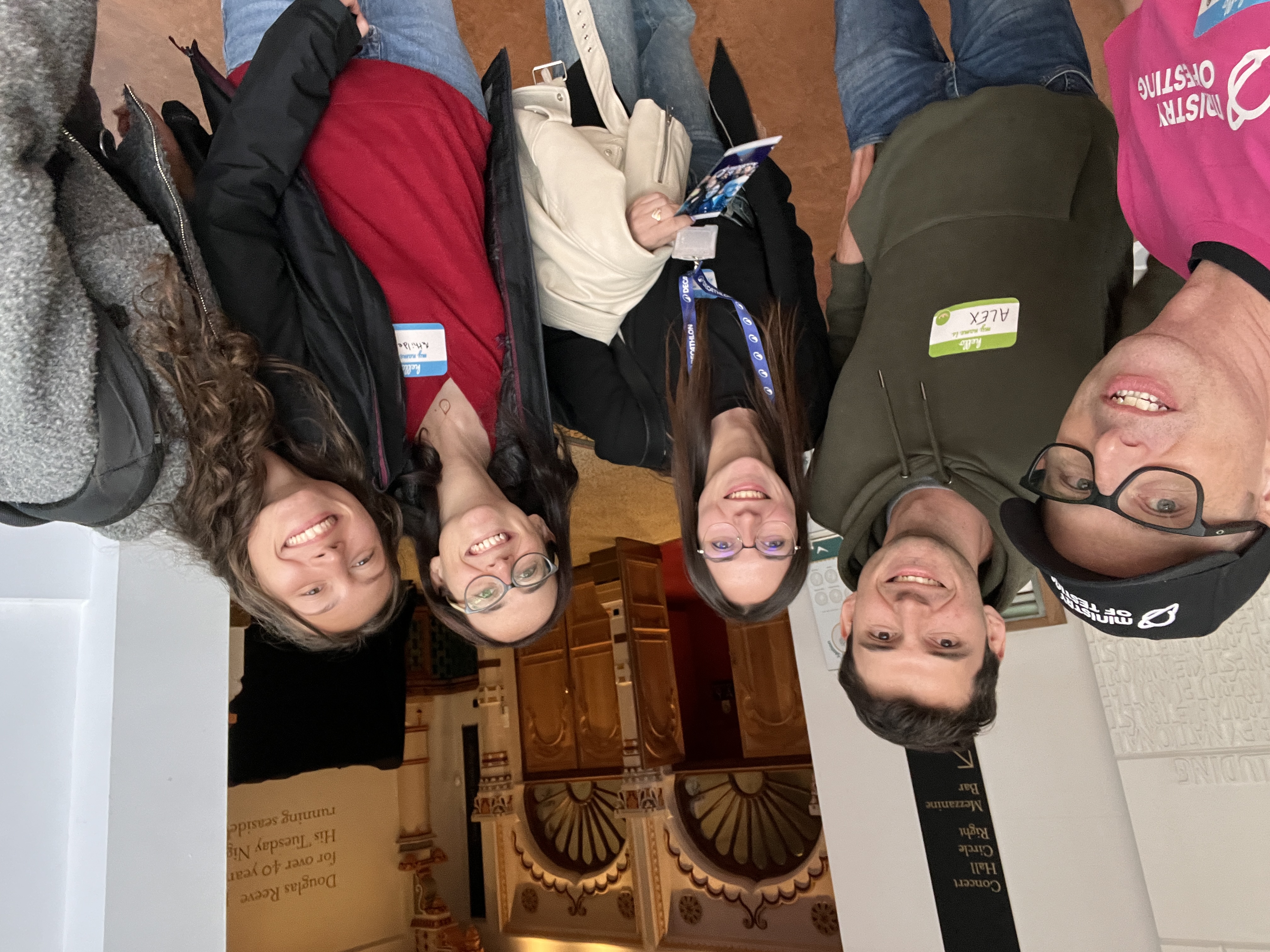 Two men and three women smile at the camera for a selfie. Some of them are wearing coloured name tags. They are standing in a grand-looking conference space with fancy-looking arches and wooden doors in the background. 