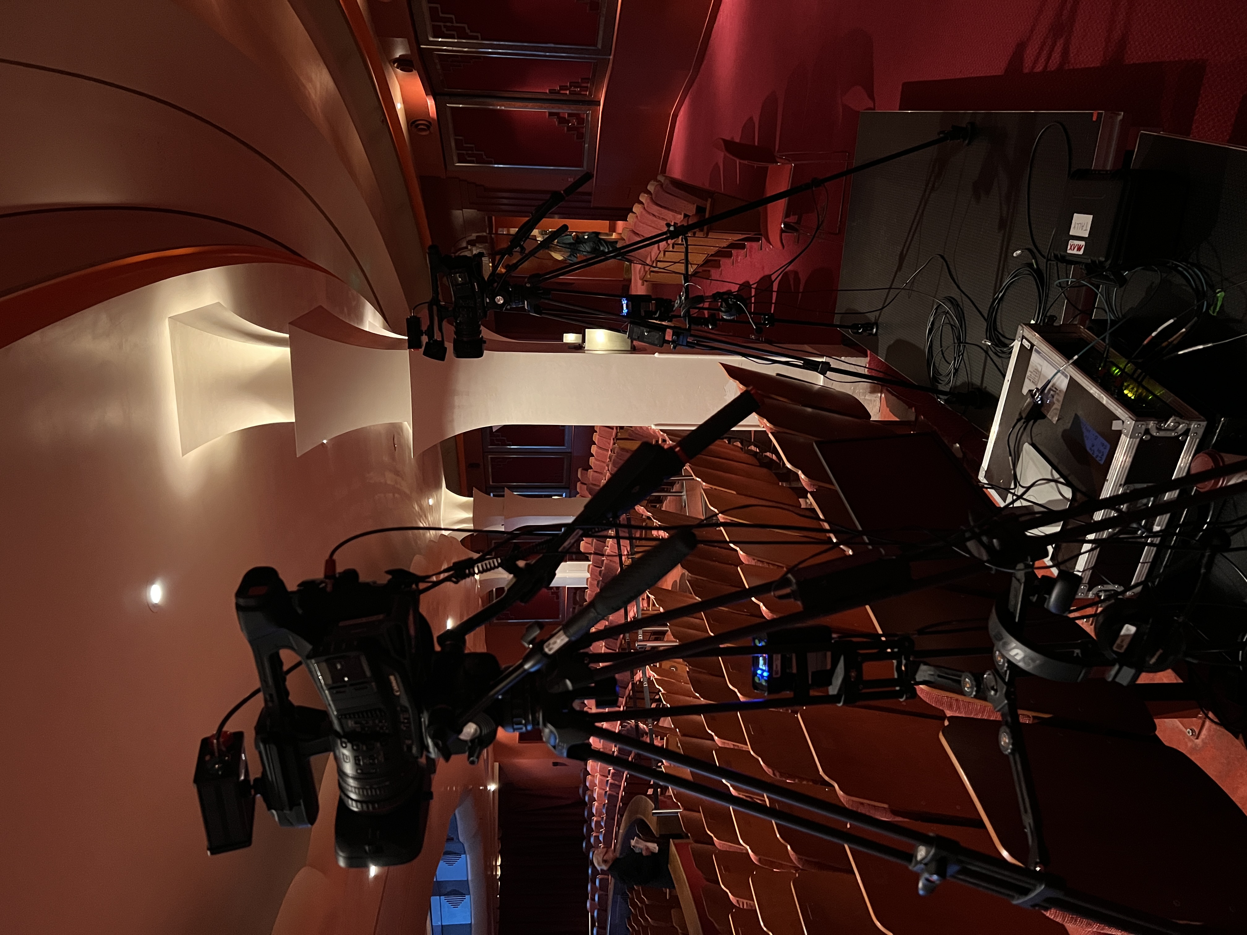 A photograph taken inside an empty theatre, looking towards the stage. Two video cameras on tripods are set up in the foreground. The theatre seats are red, and the walls are also a deep red colour, with ornate detailing and decorative columns. The lighting is dim, creating a warm, intimate atmosphere.