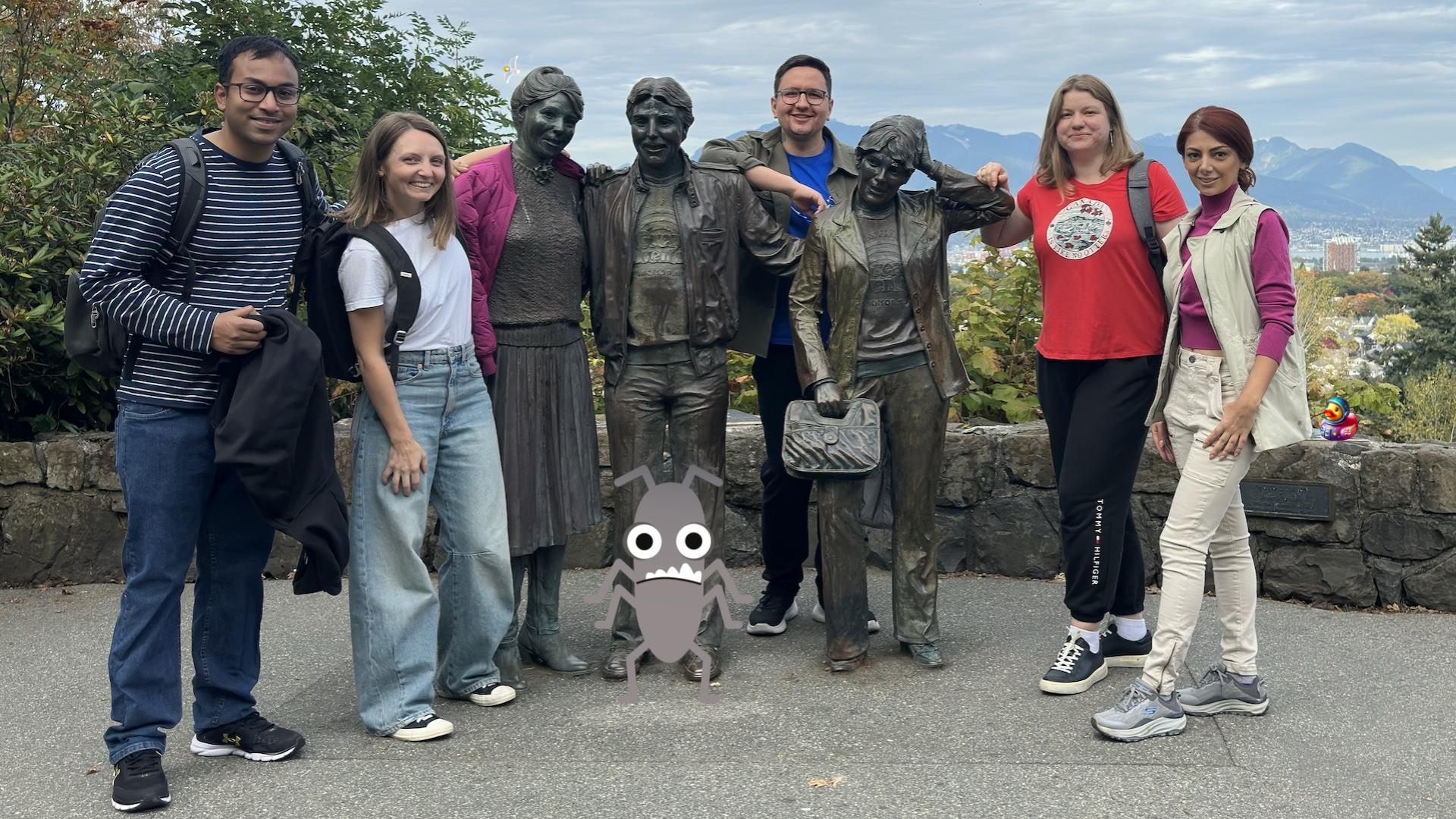 Five people stand next to three statues at the top of a hill. There are mountains in the background and clouds in the sky. A seagull, bug and duck character have been added to the photo.