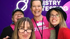 A group photo of four people at Ministry of Testing event Testbash 2024, standing in front of a purple backdrop with stars and planetary designs. Two people are wearing bright pink 'Ministry of Tes... image