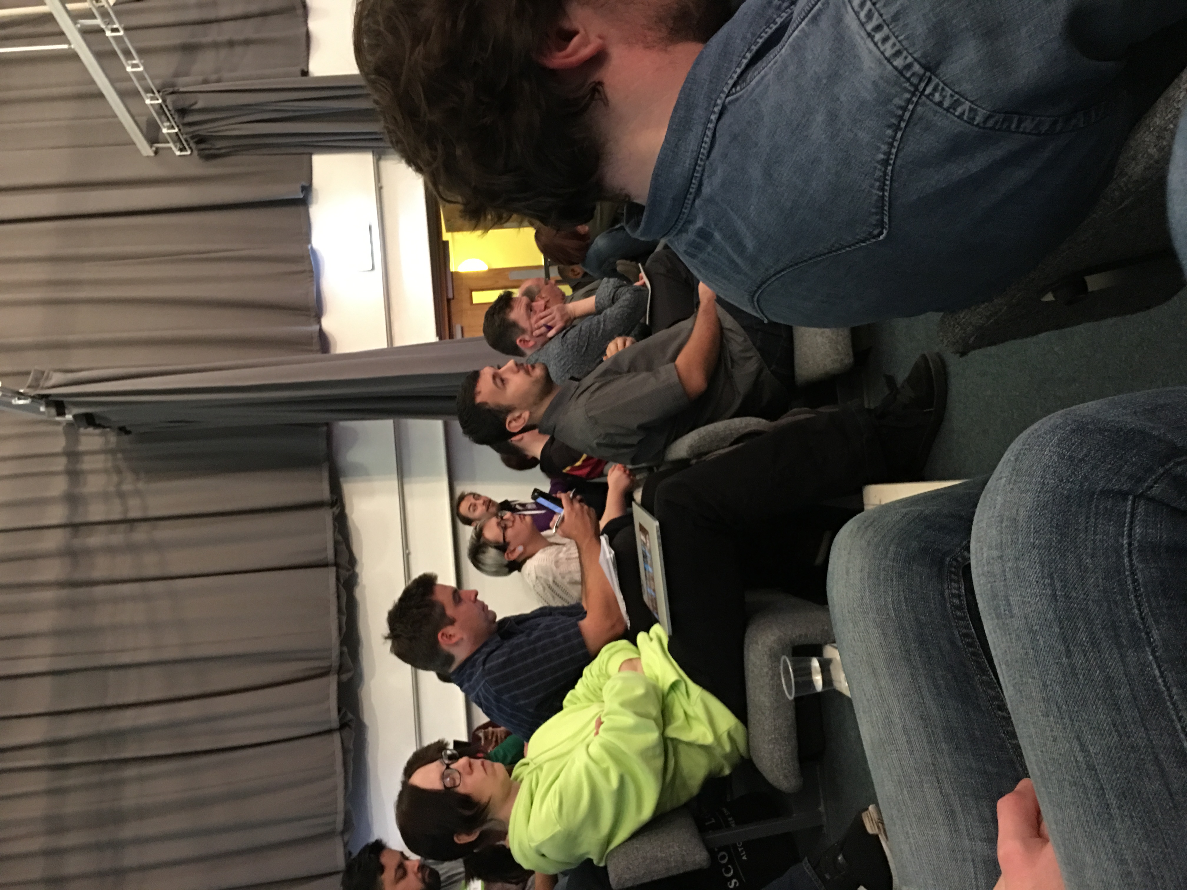 A group of people sat on auditorium-style chairs. At the end of the row is a man looking surprised. 