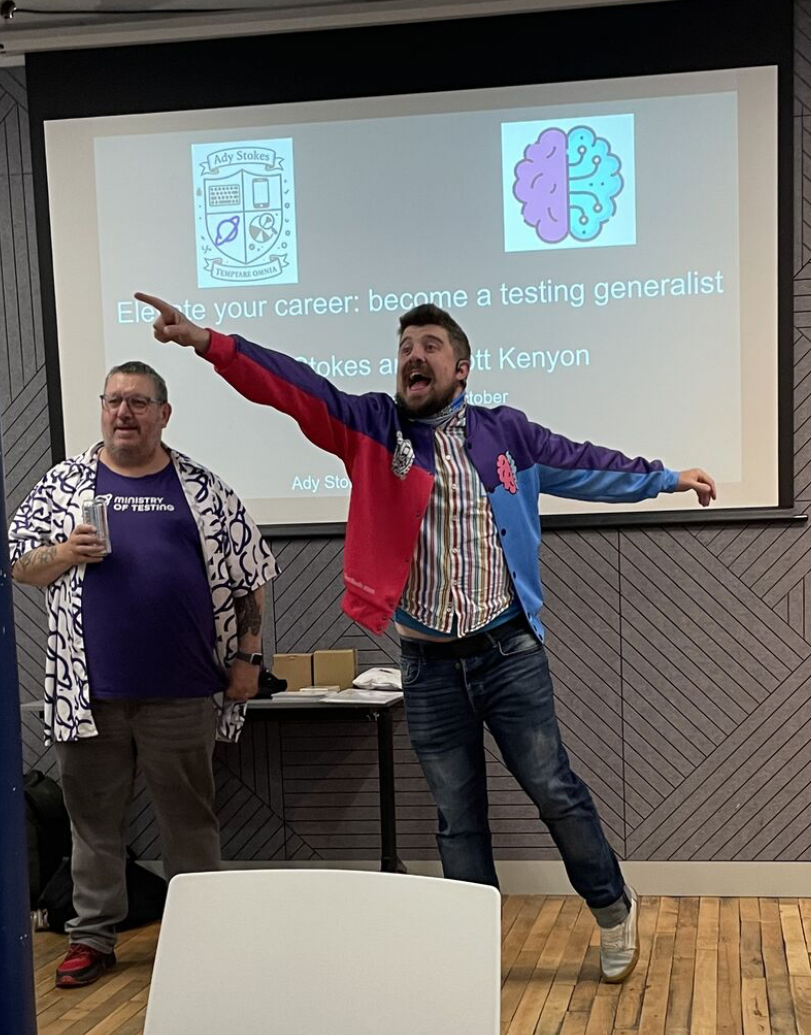 Ady Stokes and Scott Kenyon in front of a screen with their logos and workshop title. Ady has a purple MoT T-shirt and planet logo shirt over. Scott has a multi coloured jacket and is pointing into the distance for some reason only he knows. 