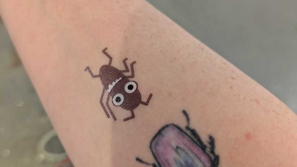 photo of a right arm with two bug tattoos: One grey and temporary one purple.
There is also a rorshach symbol on the right hand side of the wrist.
An orange dress with green frogs in various halloween costumes can also be seen.