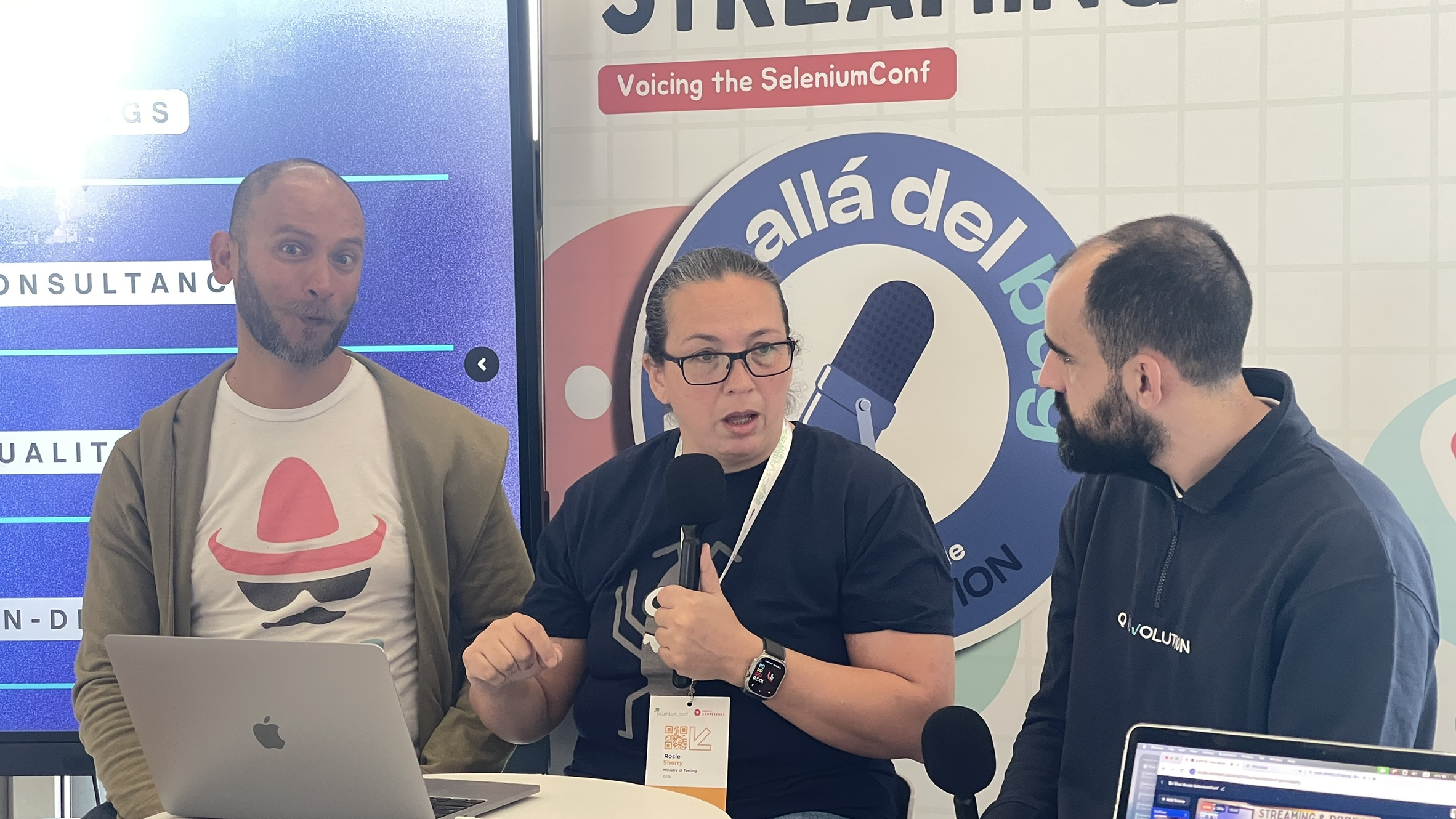 Rosie is mid-sentence, mic in hand, between two podcast hosts at the Señor Performo live podcast booth. One host is making a playful face while the other listens intently. Behind them is a big "Voicing the SeleniumConf" banner and a large mic graphic.