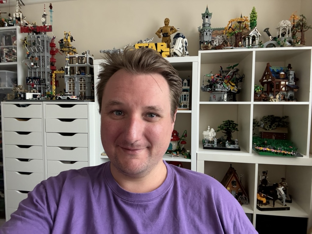 Middle aged white man with brown hair , wearing a purple t-shirt with lots lego on display in the background