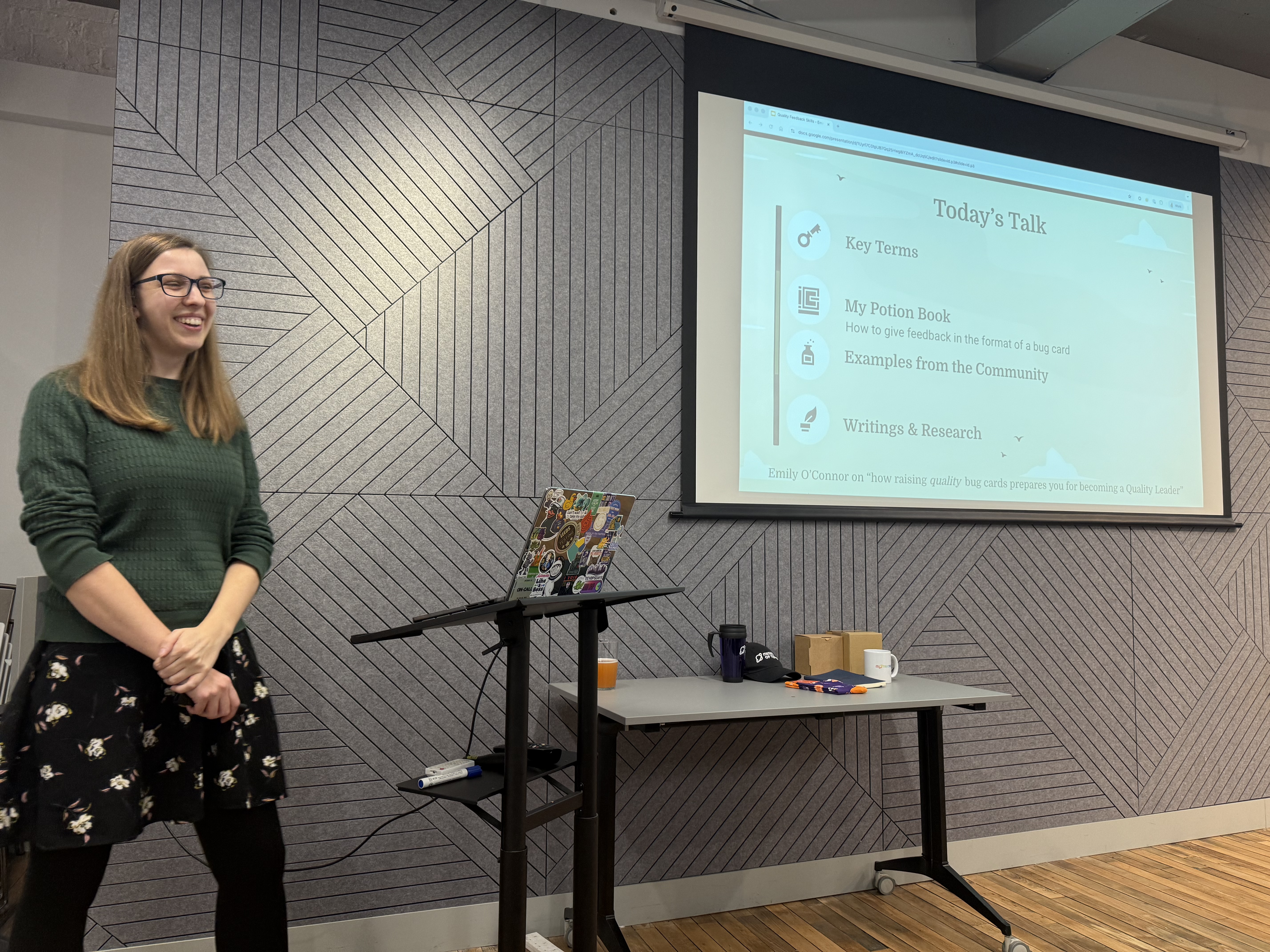 Emily stands to the side of a projector screen, wearing a green jumper and black skirt with flowers on. The screen shows a slide with today’s talk includes key terms, my potion book, how to give feedback, examples from the community, and writings and research 