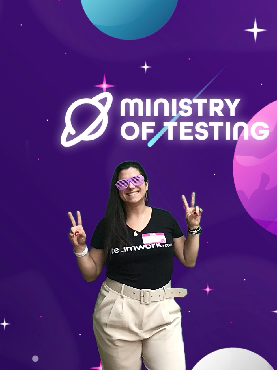 One attendee throws peace signs up in the Ministry of Testing Photobooth. They are wearing pink fun glasses and a name tag on their left. They are wearing a black tshirt that reads teamwork.com and creme trousers with a belt. They have a big smile on their face.

Background: The green screen backdrop depicts a simple illustrated outer space scene. The main colour is purple, with cute stars and planets in shades of pink, blue, and white. About a third of the way down the screen, a logo appears. It reads "Ministry of Testing" in bold white text with a blue shooting star behind it. Next to the text, in the same bold white style, is a line drawing of the planet Saturn.