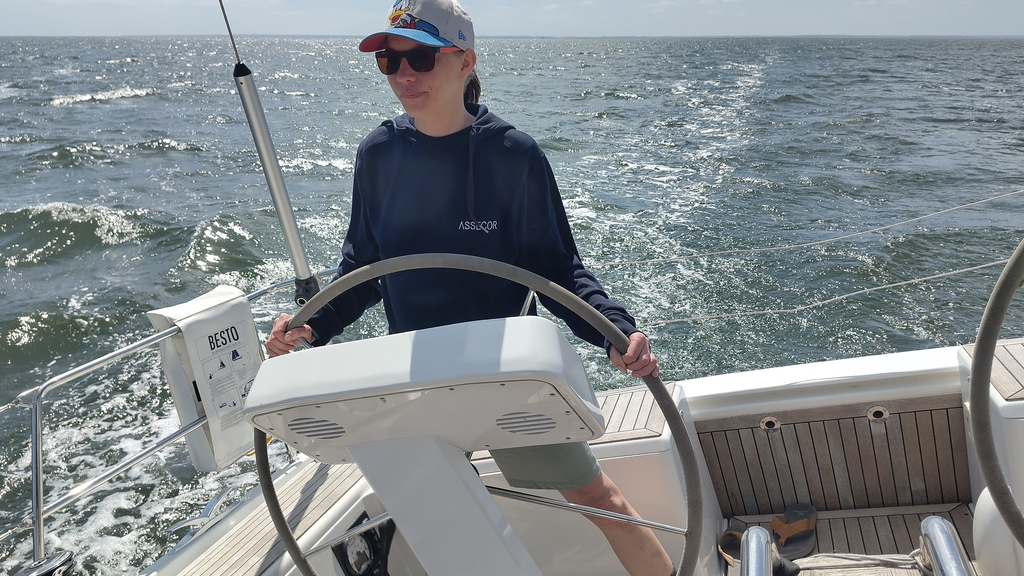 I am a younger-looking, 39-year old, white female, wearing beige bermuda shorts, a blue hoodie with the company name (Assecor) in white, a Donald Duck basecap and orange sunglasses. I am holding the steering weel of a 47ft-long yacht, behind me the horizon of the Bay of Greifswald (that's just lots of water), the tilted ship is indicating that we're sailing.