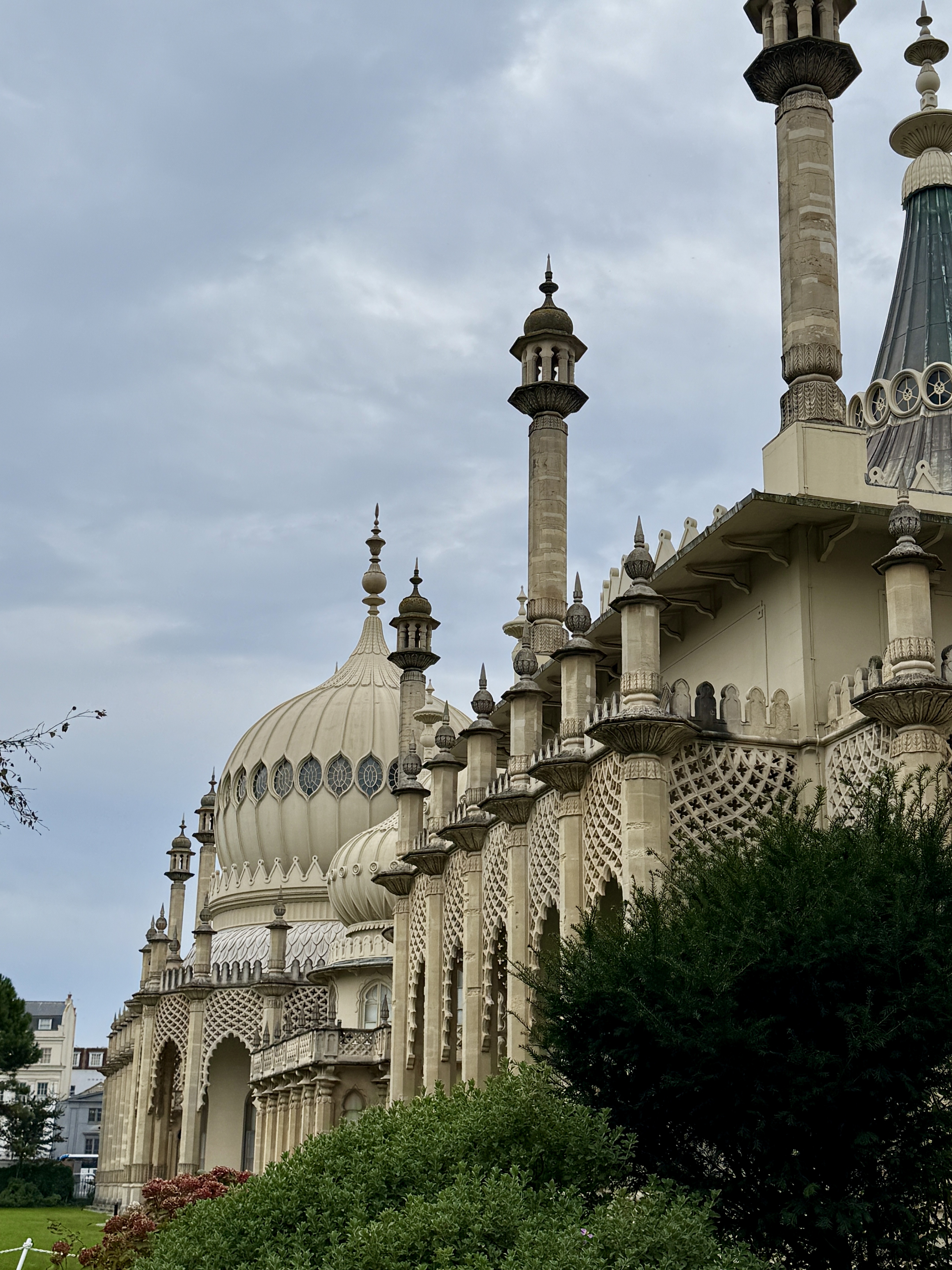 Low-angle shot of the Brighton Pavilion's elaborate Indo-Saracenic architecture, featuring minaret-like towers, a large onion dome with arched windows, intricate stone tracery, and dark green foliage in the foreground, all under a cloudy sky.