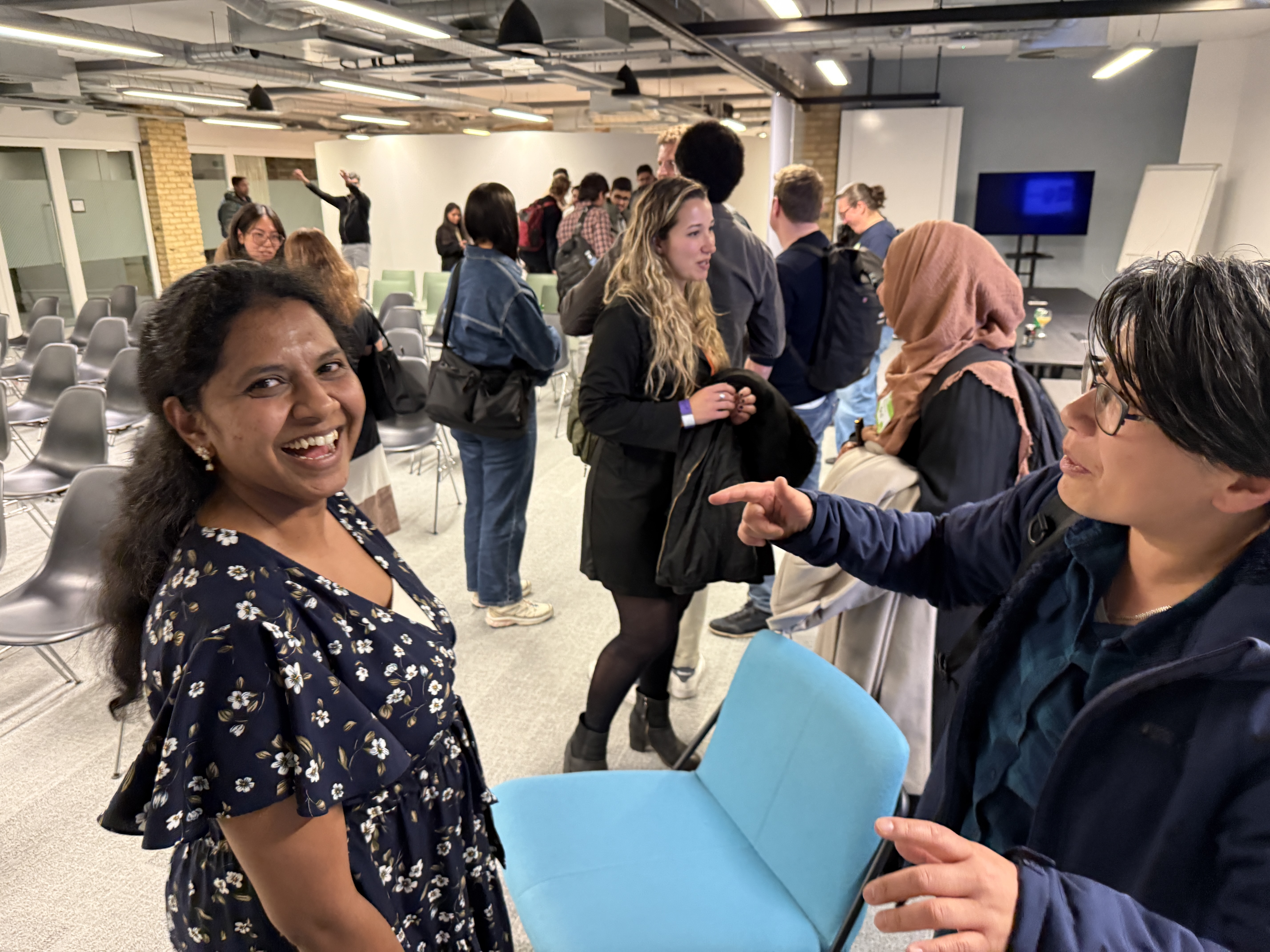 One person smiles at the camera whilst another person gestures towards them. In the background, people are mingling in an office event space.