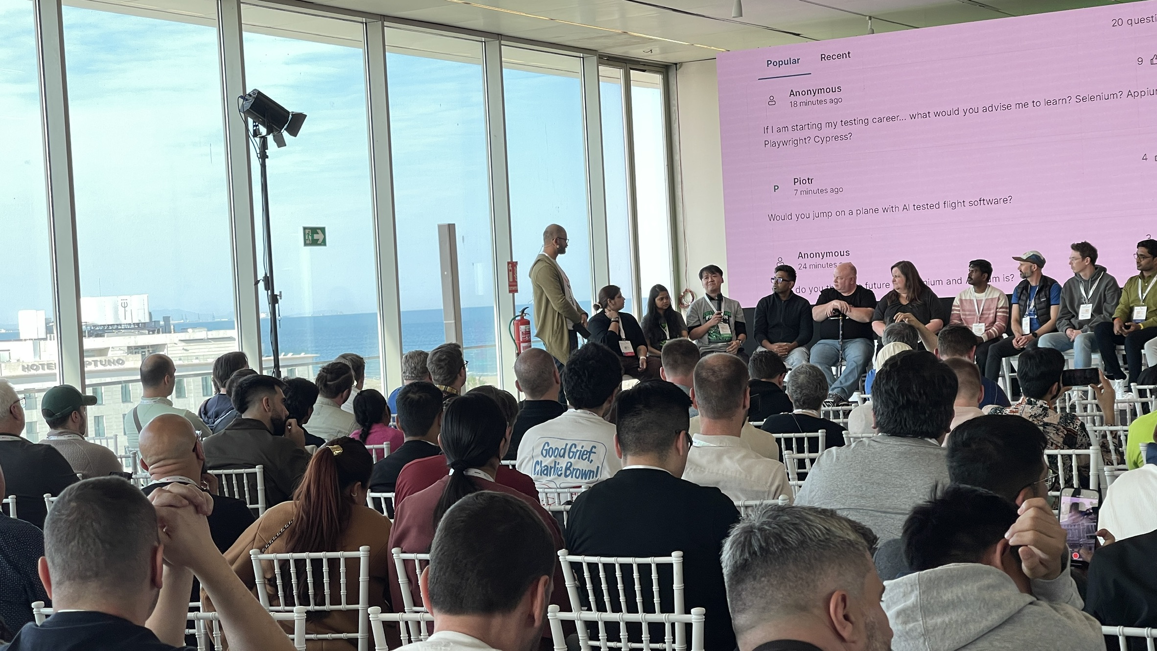 A large panel of speakers faces a packed audience at the closing session of SeleniumConf 2025. The backdrop displays live Q&amp;A questions, with the ocean visible through the venue’s panoramic windows. Host Leandro stands to the left, mic in hand, guiding the final reflections.