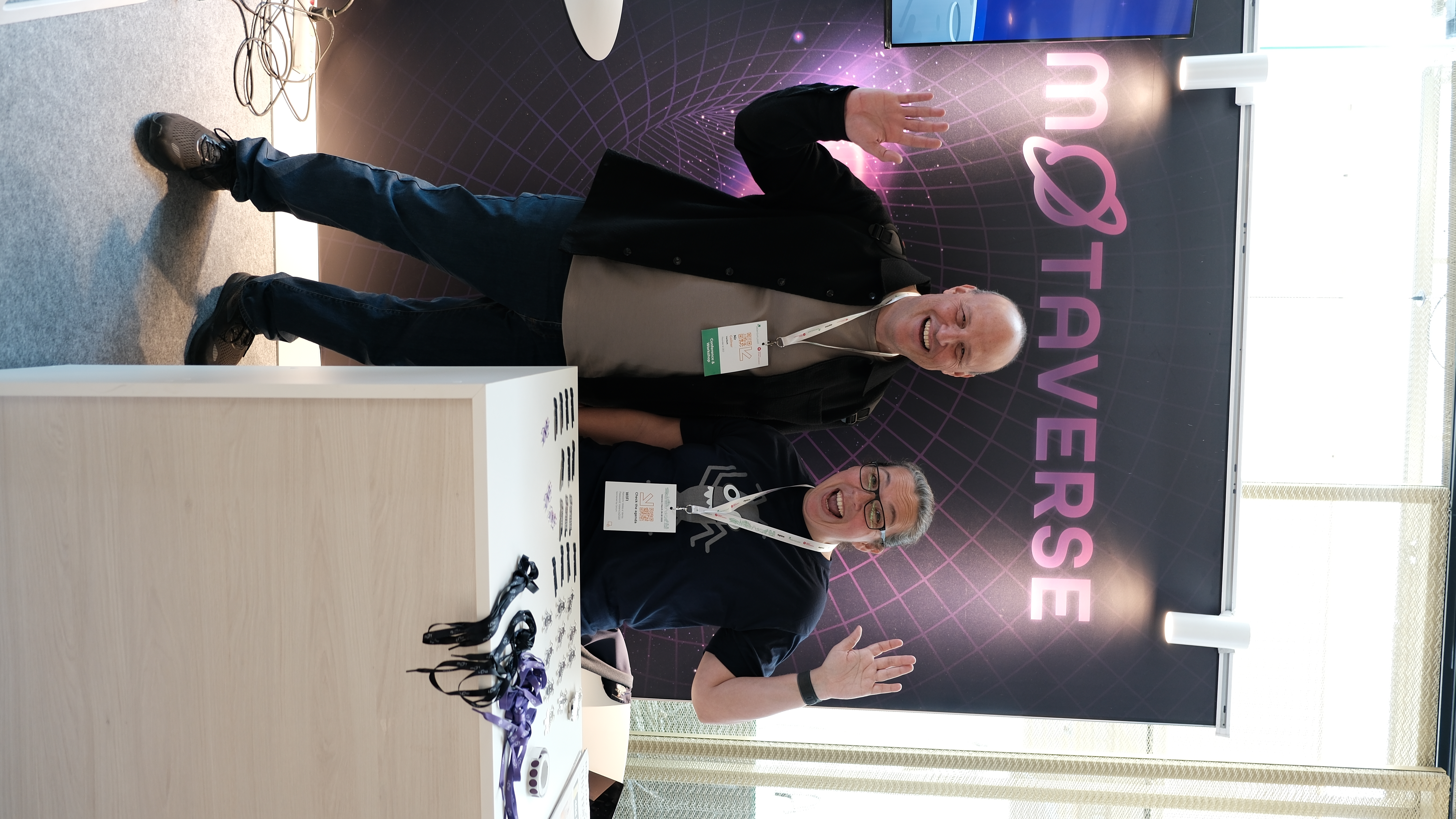 Nir Gallner and Rosie Sherry stand smiling and waving at the Ministry of Testing stand during Selenium Conf 2025, in front of the MoTaverse backdrop, with stickers and laces on the table in front of them.