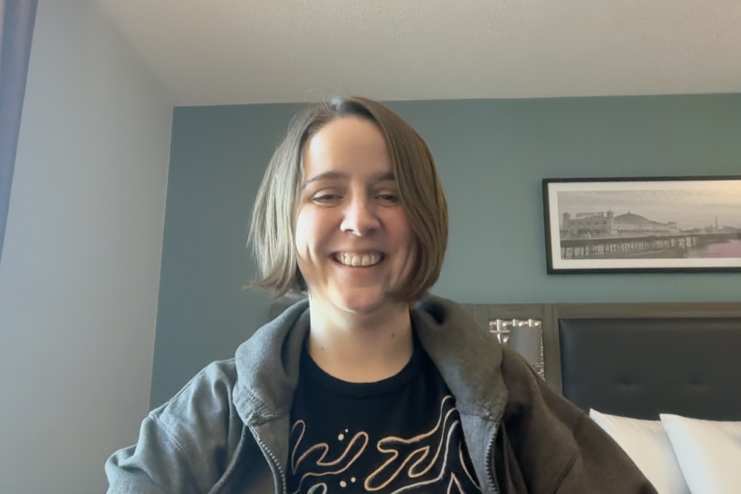 Cheery selfie from a middle aged woman excited for test bash. She is wearing a grey hoodie and is sat at a desk in a hotel room.