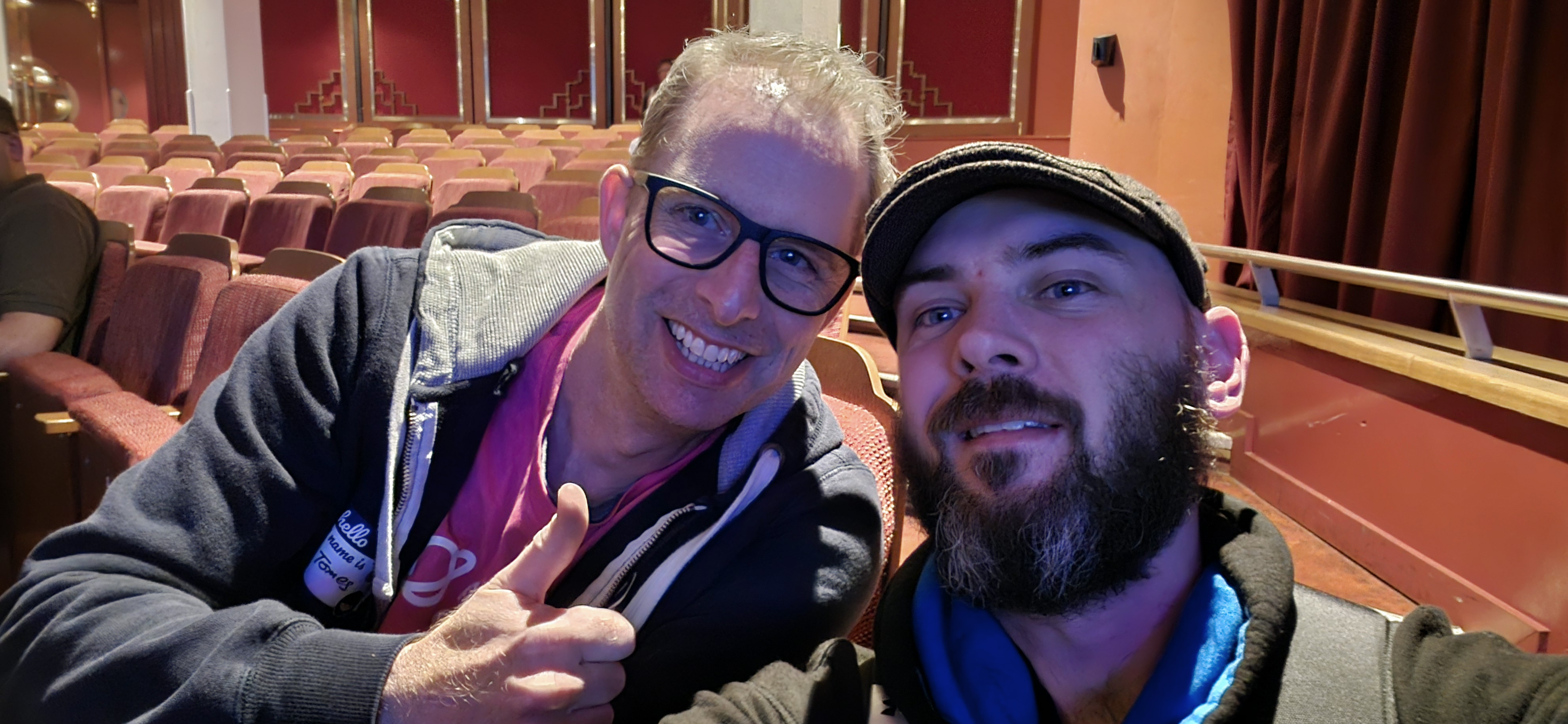 Two people smile for a selfie in a theatre with red seats and decorative walls. One person gives a thumbs up, wearing glasses and a hoodie. The other person wears a cap and has a beard.