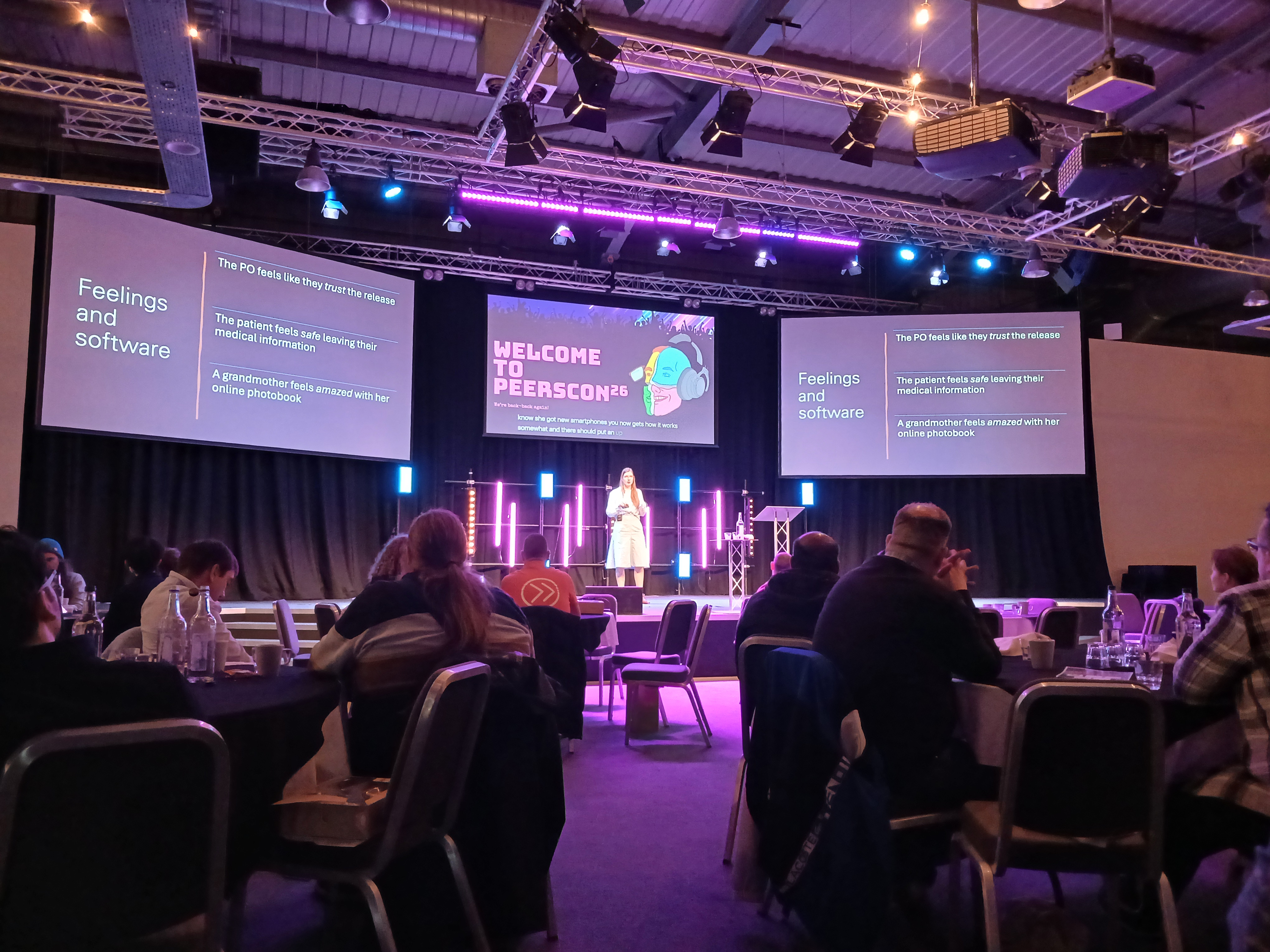 Woman delivering a talk at Peerscon on stage with 3 screens, 1 behind her show the conference titles and 2 others showing the slides of her presentation to the entire room