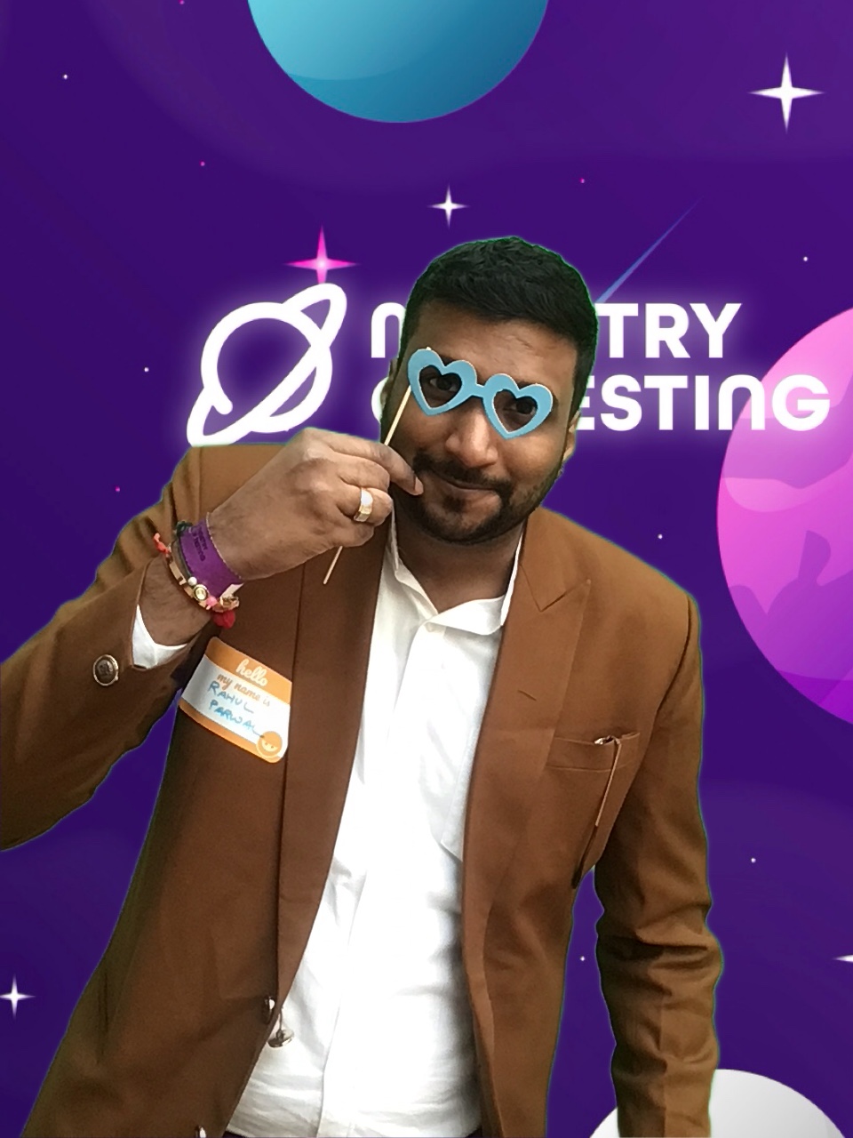 This attendee is wearing a brown suit with a white shirt and a name tag on his right side. The most striking part of this image has to be the blue heart shaped glasses on a stick held to their eyes to look straight through and into the camera with a smile. 

Background: The green screen backdrop depicts a simple illustrated outer space scene. The main colour is purple, with simple twinkly stars and planets in shades of pink, blue, and white. About a third of the way down the screen, a logo appears. It reads "Ministry of Testing" in bold white text with a blue shooting star behind it. Next to the text, in the same bold white style, is a line drawing of the planet Saturn.