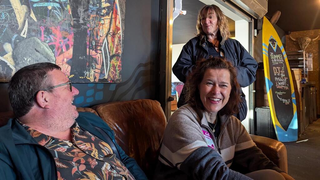 
A medium shot captures three adults in what appears to be the inside of a pub/bar. In the foreground, a man with glasses and a patterned shirt is seated on the left, looking off-camera to the left with a smile or laugh. Next to him, a woman with dark hair and a striped sweater is seated, smiling broadly directly at the viewer. In the background, another woman with lighter, curly hair and a dark jacket stands in a doorway, looking towards the man. The setting features dark walls, some vibrant abstract art, and a yellow surfboard leaning against a wall in the background. A bug, a seagull and a duck character have been added to the photo. 