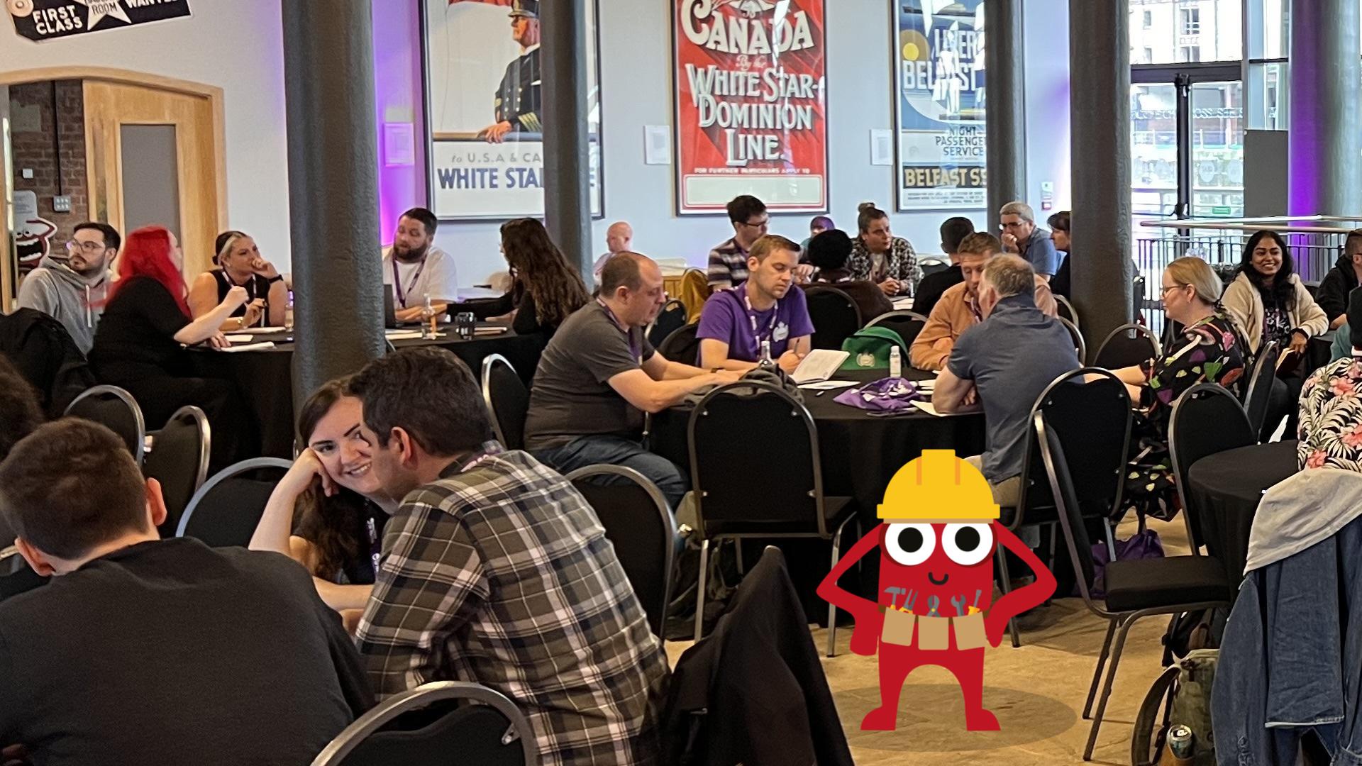 A workshop setting with groups of people gathered around circular tables discussing testing topics. A monster character with a builders hat is in the foreground looking directly at the camera.
