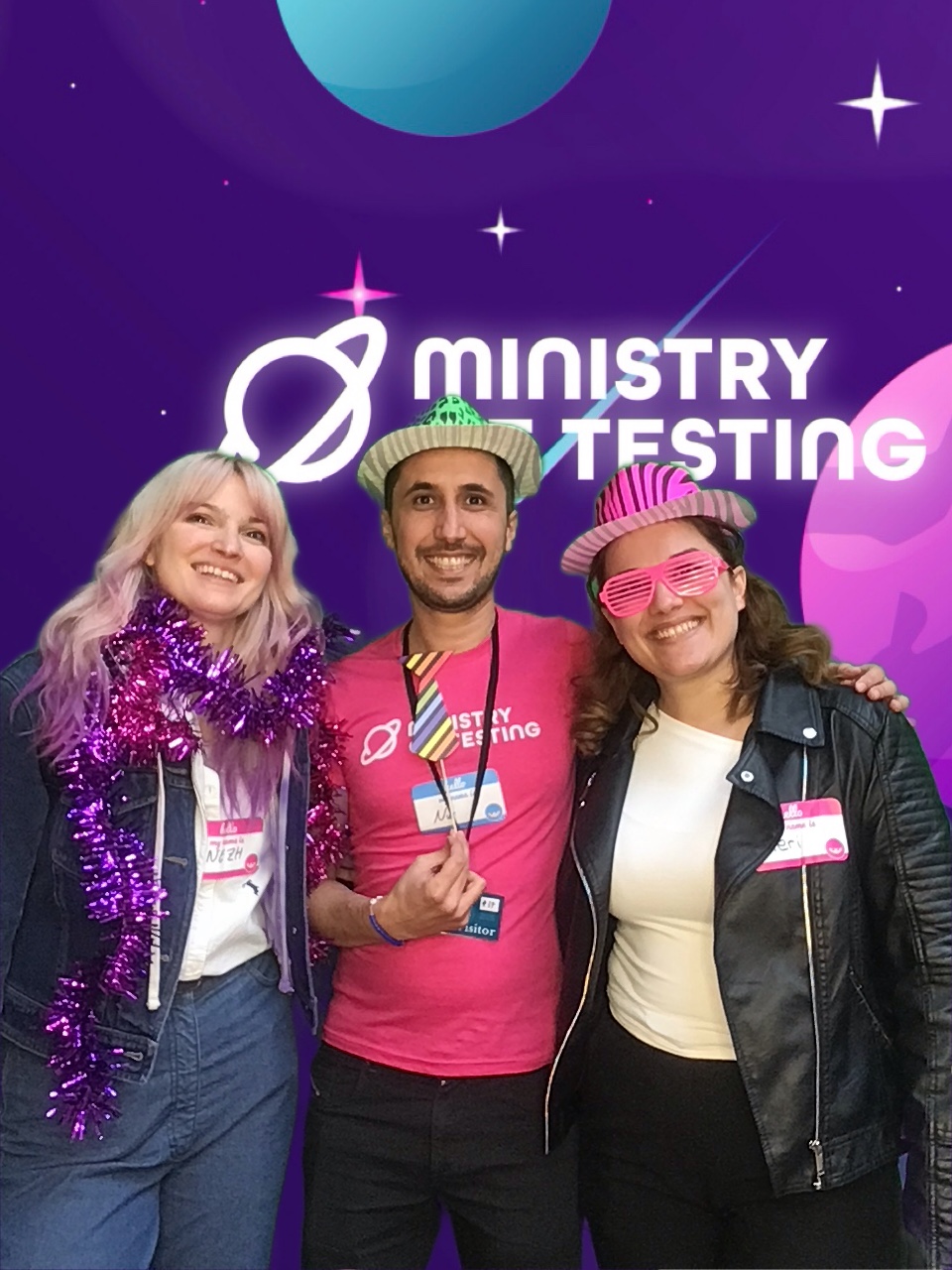 A happy trio donned in a variety of fun and festive accessories. Smiles all round. 

Attendee 1 (on the left) has blonde hair with purple tips and purple sparkly tinsel around their neck. 

Attendee 2 (middle) has a fun green leopard print hat, and a rainbow tie on a stick that they are holding in front of their necks like a real tie. They also are wearing a pink Ministy of testing tshirt. 

Attendee 3 wears a pink zebra hat with matching pink glasess. They also have on a black leather jacket with a white top underneath. 