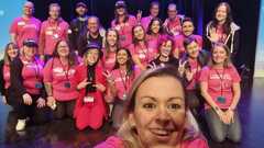 A group of several people kneeling and standing up on a conference stage. There is a woman at the front taking a selfie. Most people wear pink t-shirts with a Ministry of Testing logo. Most people ... image