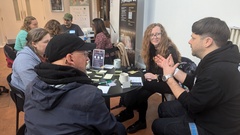A group of people participating in a Lean Coffee session at TestBash 2024. They are all seated around small round tables, engaged in discussion and exchanging ideas related to software testing. Sti... image